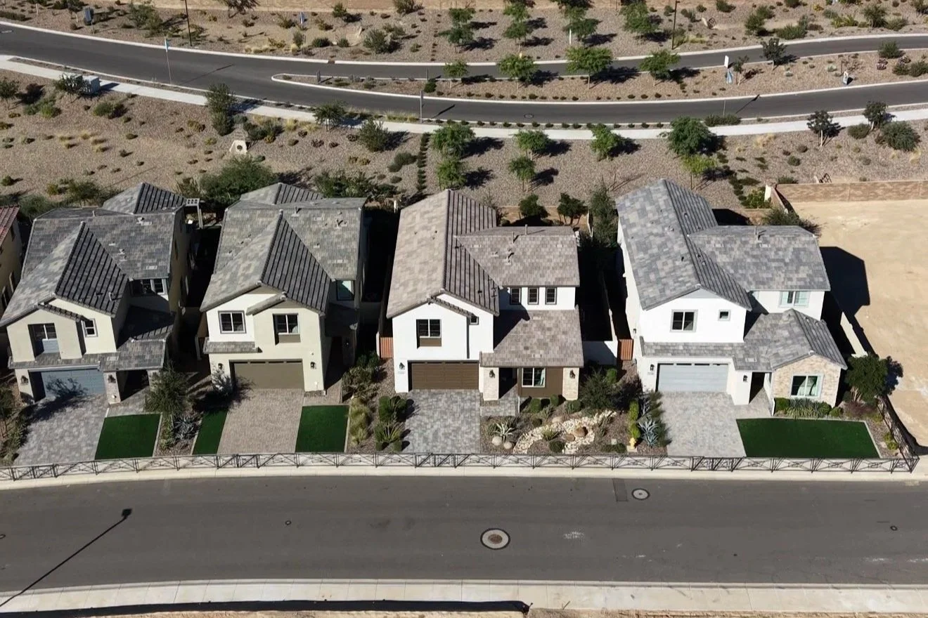 Aerial view of four newly built suburban houses with front yards, driveways, and a fence, located along a paved street with a park or pathway behind them.
