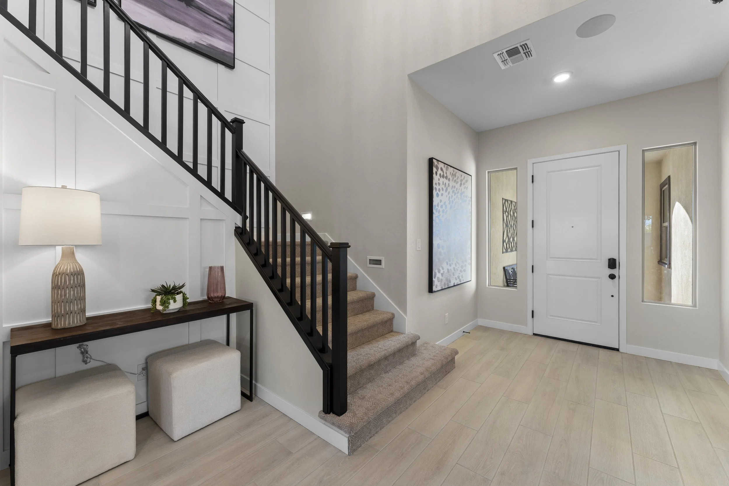 Modern front entryway with white walls, light wood flooring, a front door with side windows, a staircase with black railing, artwork on the wall, and a console table with a lamp, plant, and decorative items.