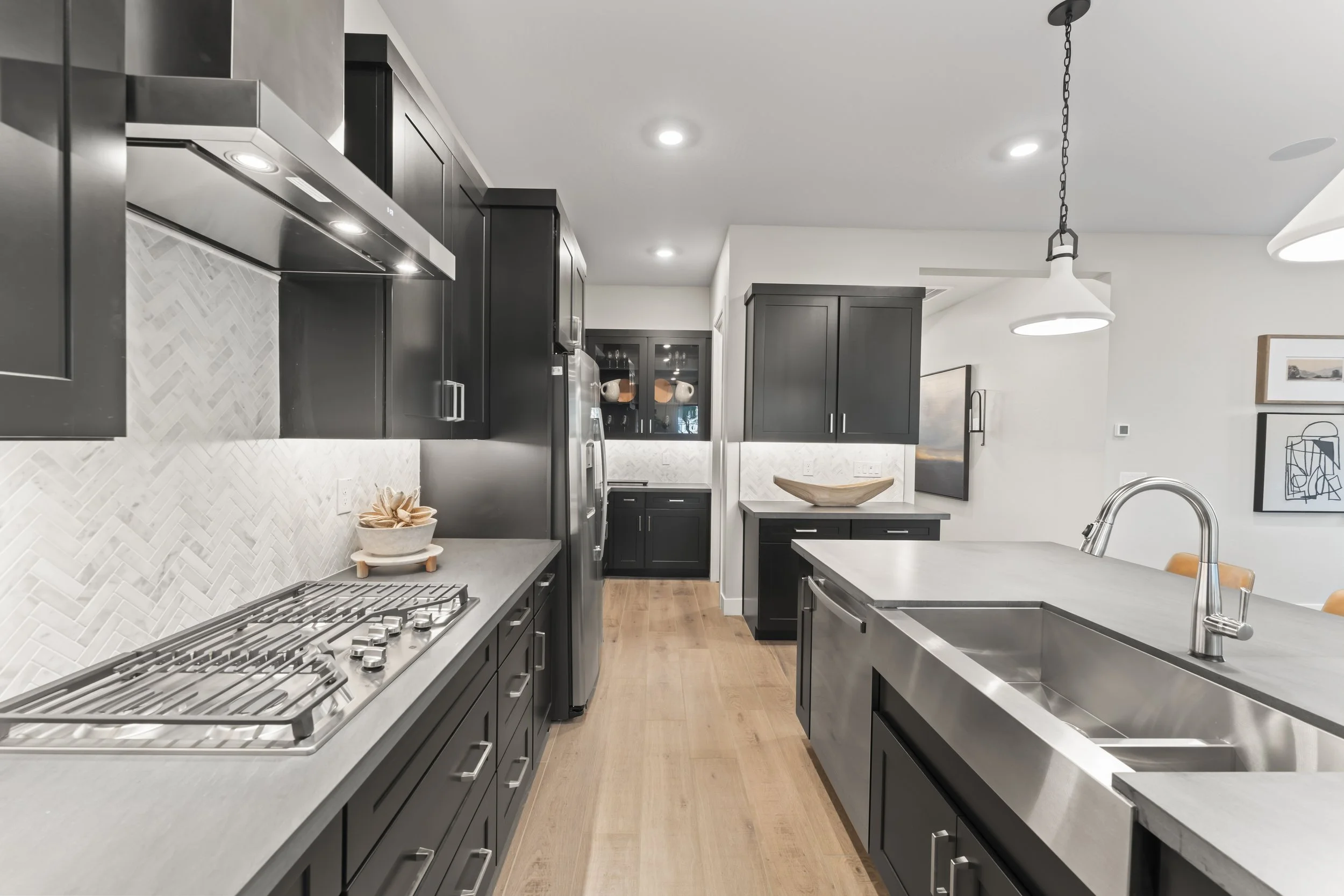 Modern kitchen with black cabinets, stainless steel appliances, white herringbone backsplash, light wood flooring, and minimalist decor, including a large kitchen island with a sink and two pendant lights.