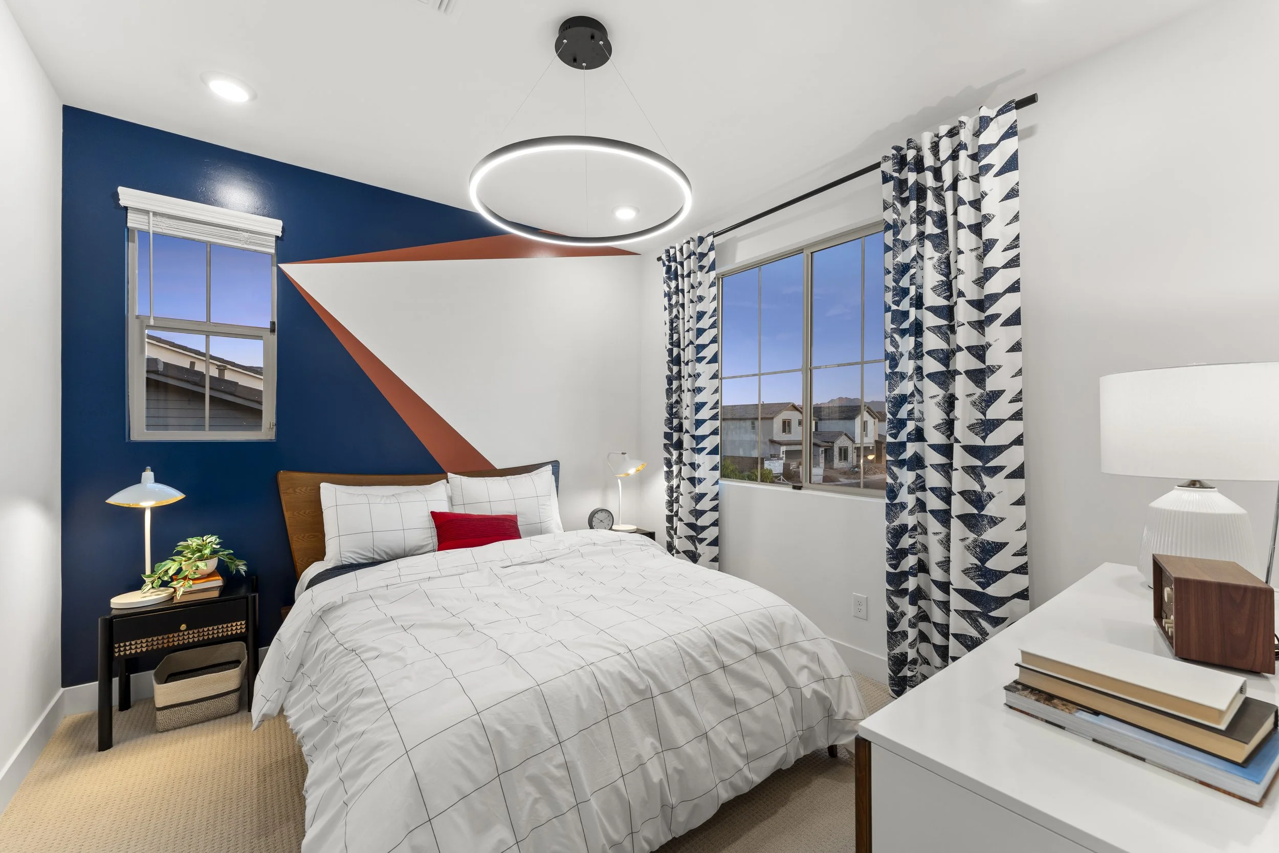 Modern bedroom with a white bed, navy accent wall with geometric design, large window with patterned curtains, bedside table with a lamp and plant, dresser with books and lamp, geometric ceiling light.
