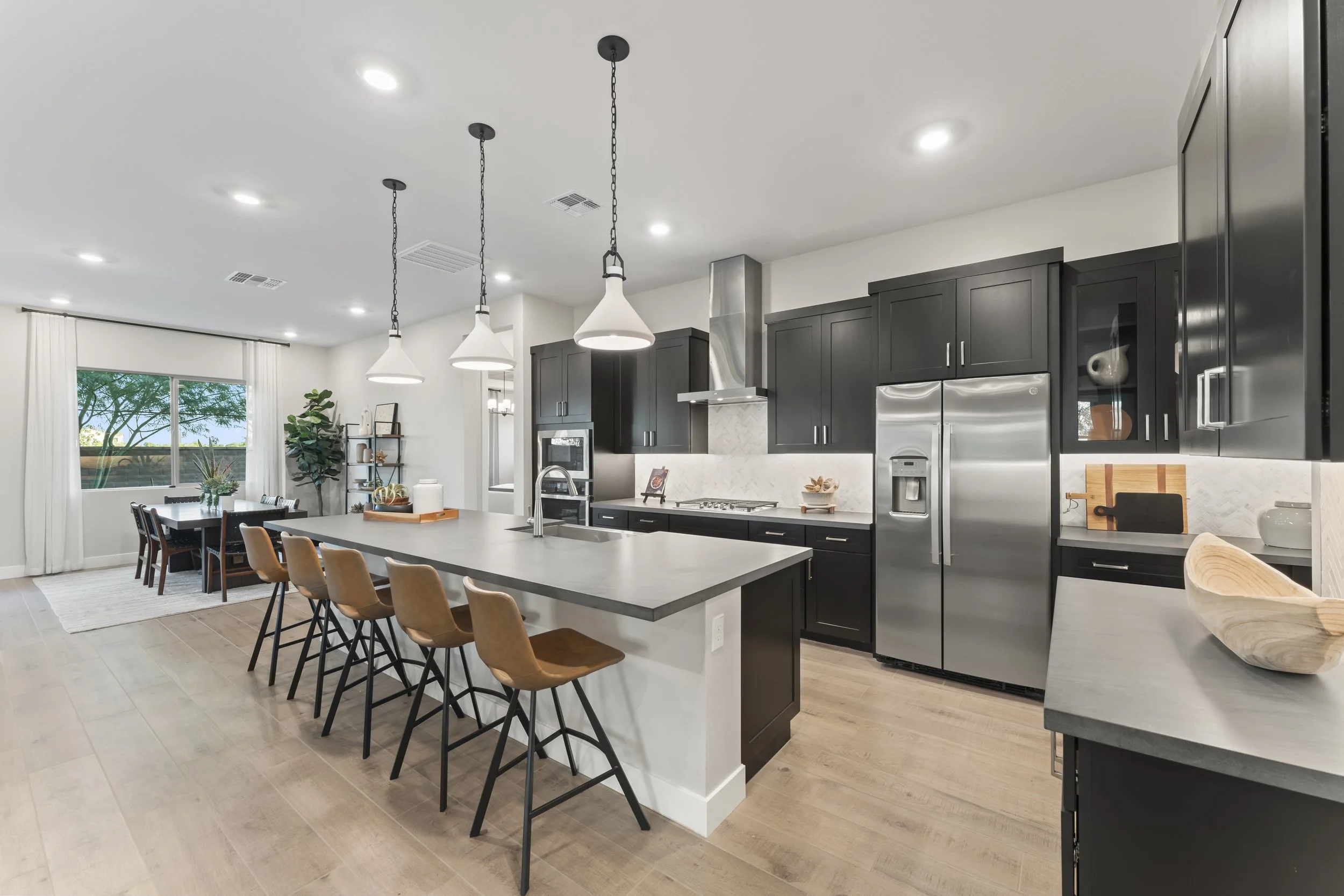 Modern kitchen with black cabinets, stainless steel refrigerator, gray island with bar stools, dining area with window, and light wood flooring.