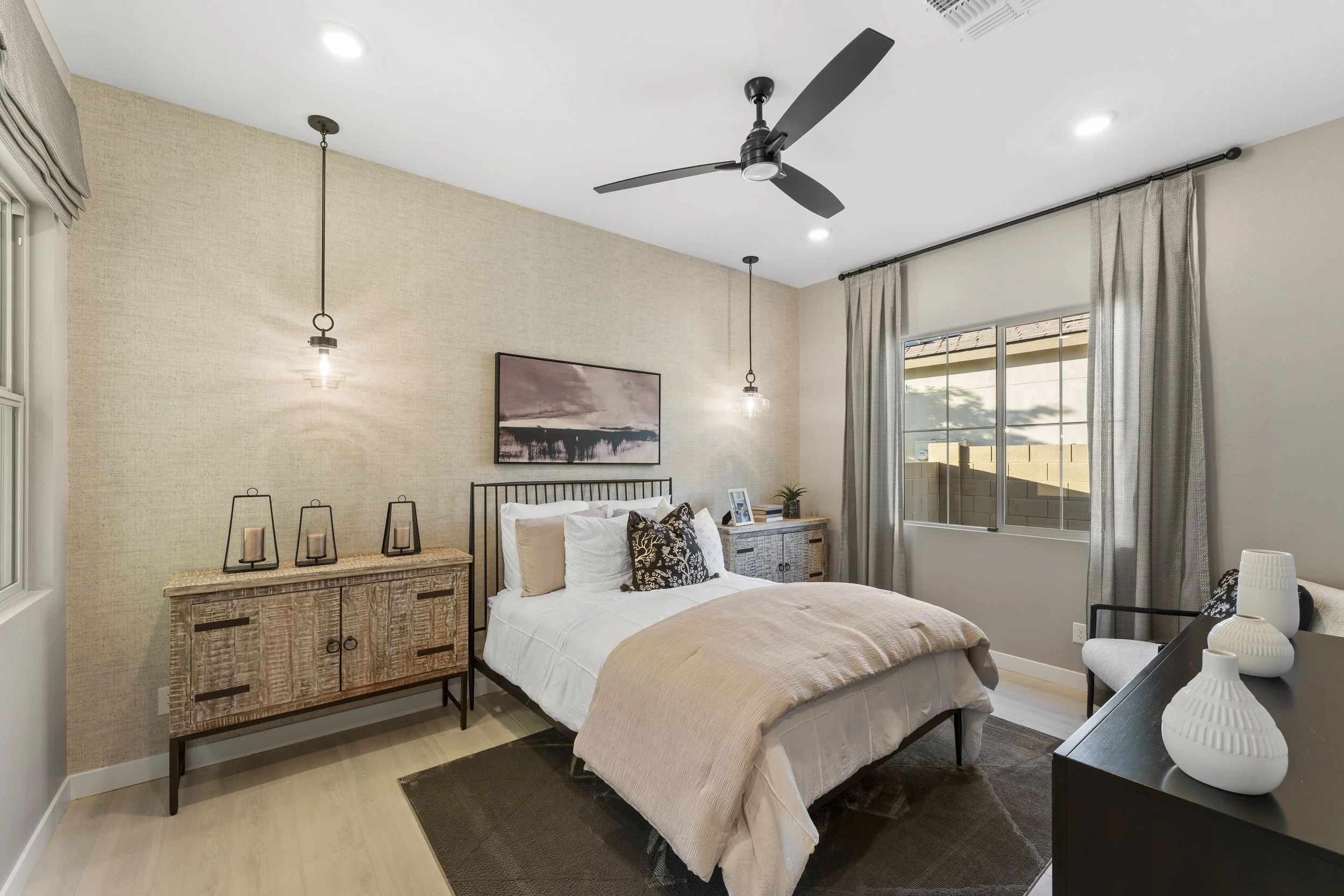 A cozy bedroom with a bed, nightstands, and decorative items. The bed has white and beige bedding with black and patterned pillows. There are two pendant lights hanging on either side of the bed, a large window with beige curtains, a ceiling fan, and