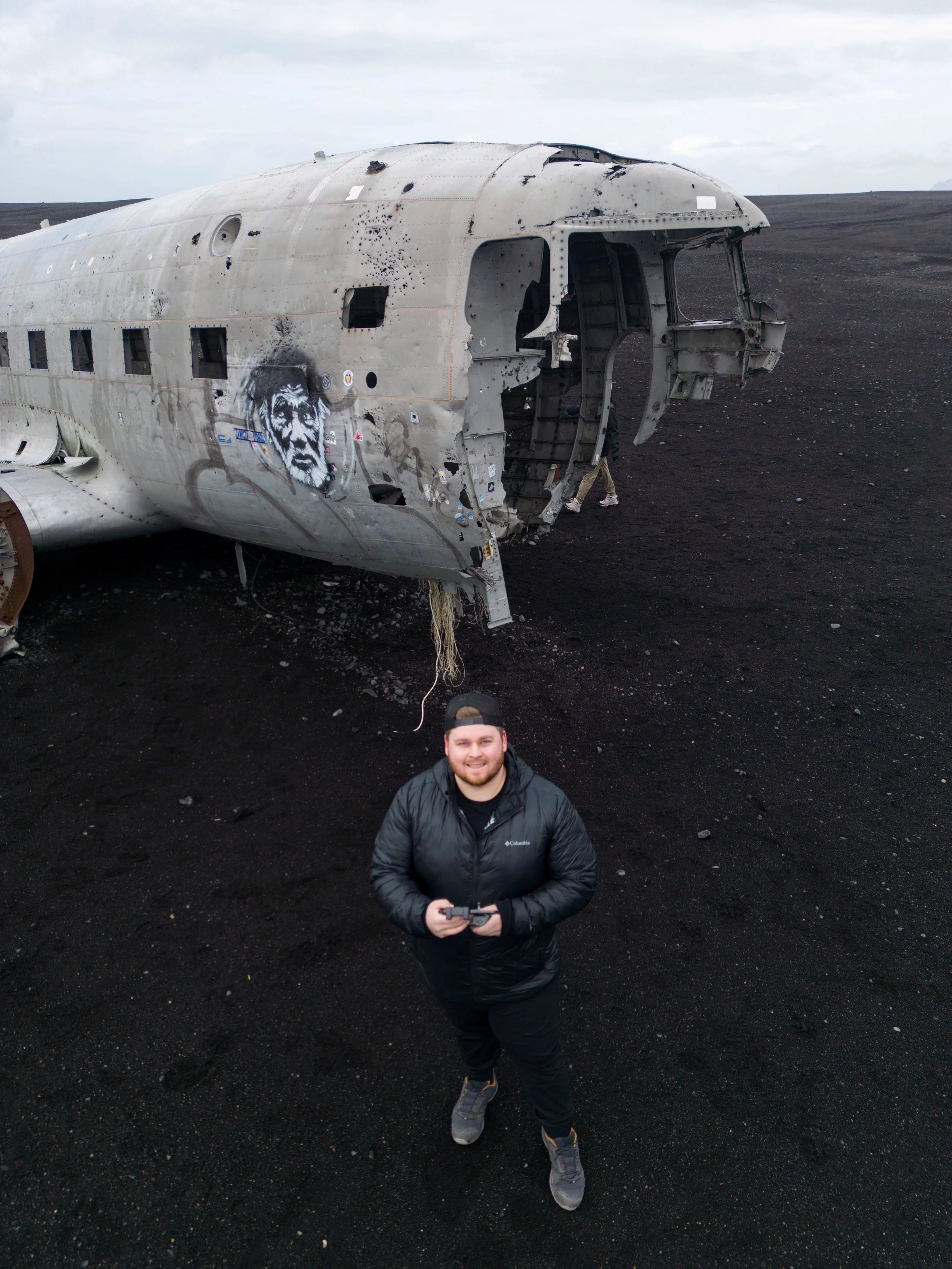A man standing on black volcanic sand in front of a wrecked airplane, holding a remote control, with a face graffiti on the airplane's fuselage.
