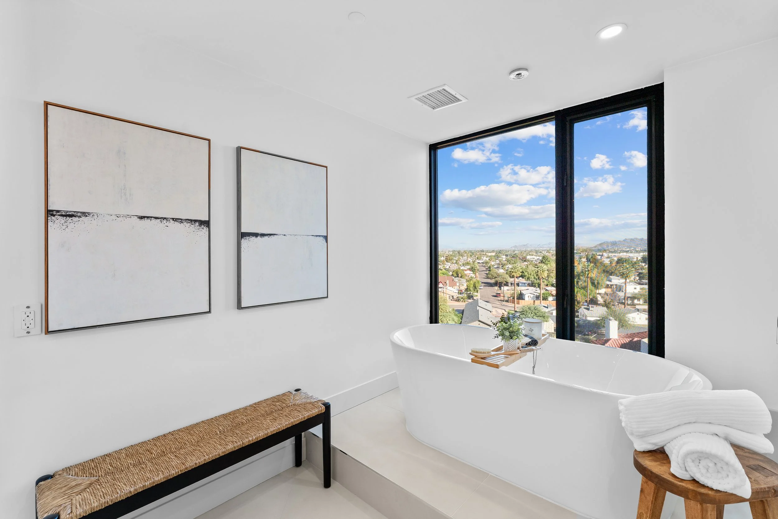 Bright modern bathroom with a white bathtub near large window showing a cityscape and blue sky with clouds, featuring minimal decor, wall art, and a bench with a woven seat.