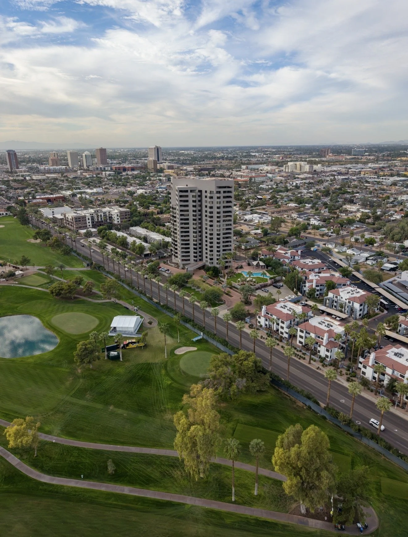 Aerial view of a cityscape showing a mix of residential buildings, a golf course with green fairways, a small pond, and tall palm trees, with a cloudy sky overhead.