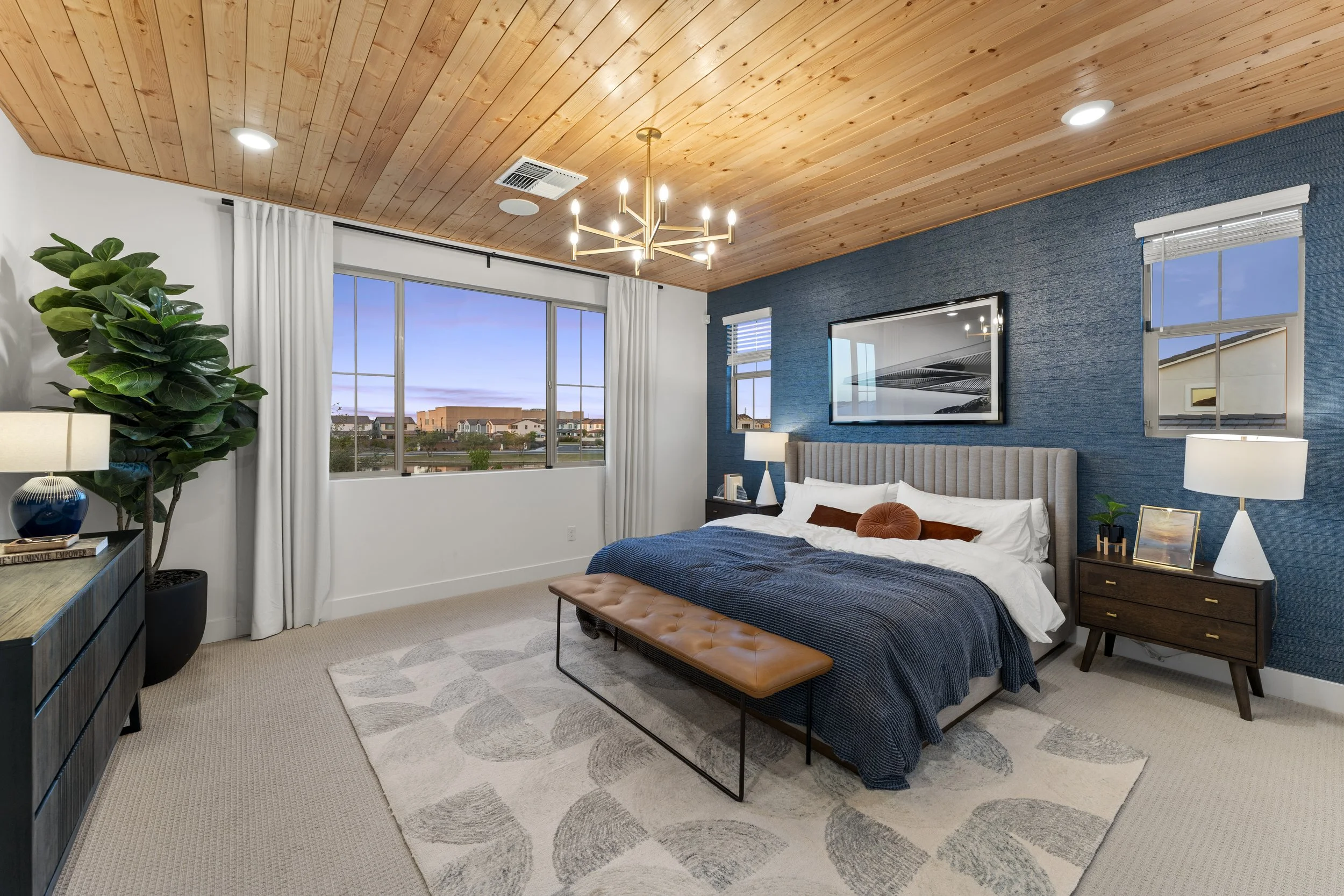 A modern bedroom with a blue accent wall, beige upholstered headboard, and a large window showing a cityscape view. The room features a leather bench at the foot of the bed, two nightstands with lamps, and a potted plant in the corner. The ceiling is