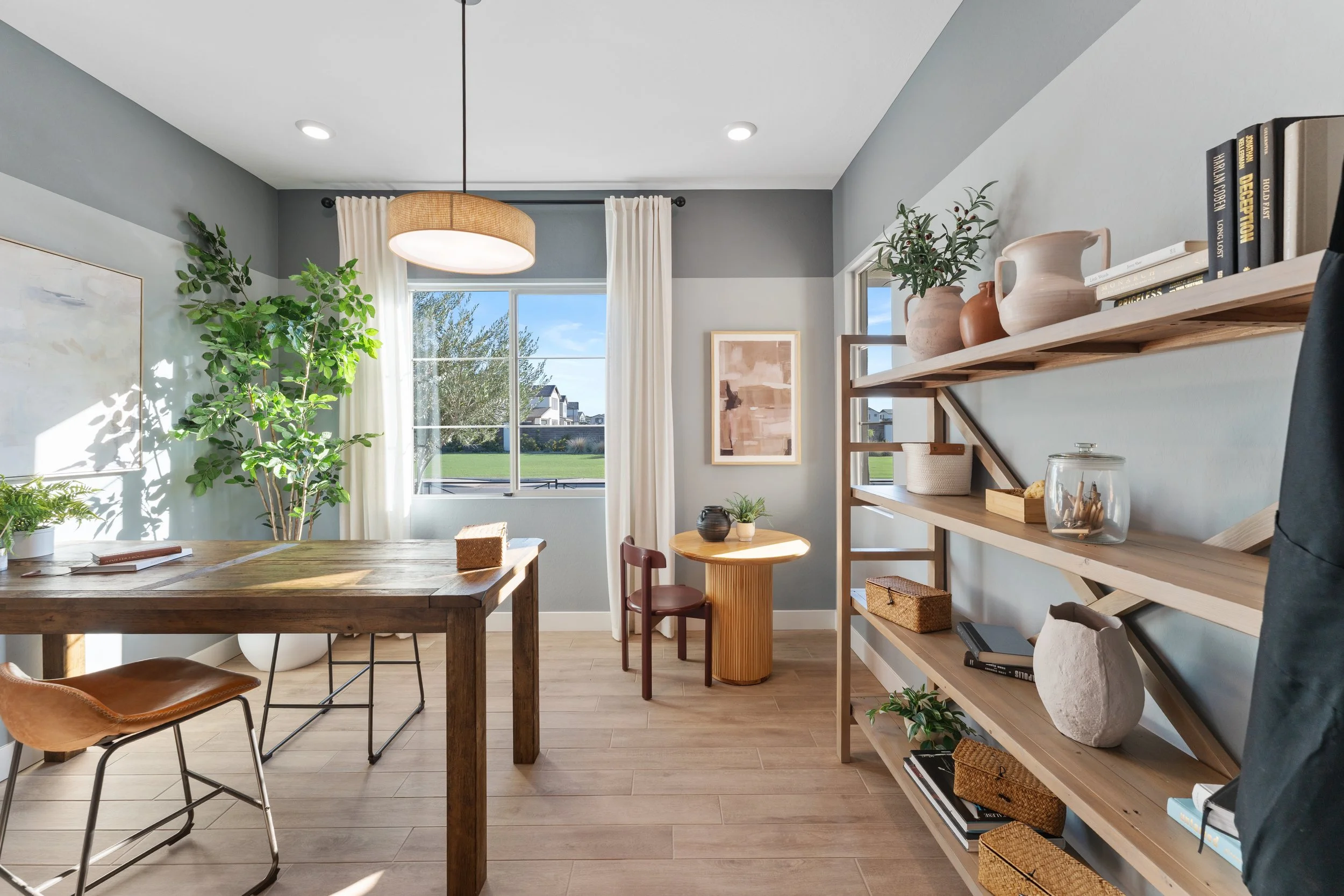 Bright living room with large window, light blue walls, wooden furniture, potted plants, and decorative objects on shelves.