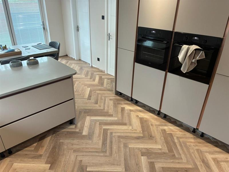 Modern kitchen with wooden herringbone floor, white cabinets, and built-in black ovens. A kitchen island with bowls is visible, along with a small dining area near a window.