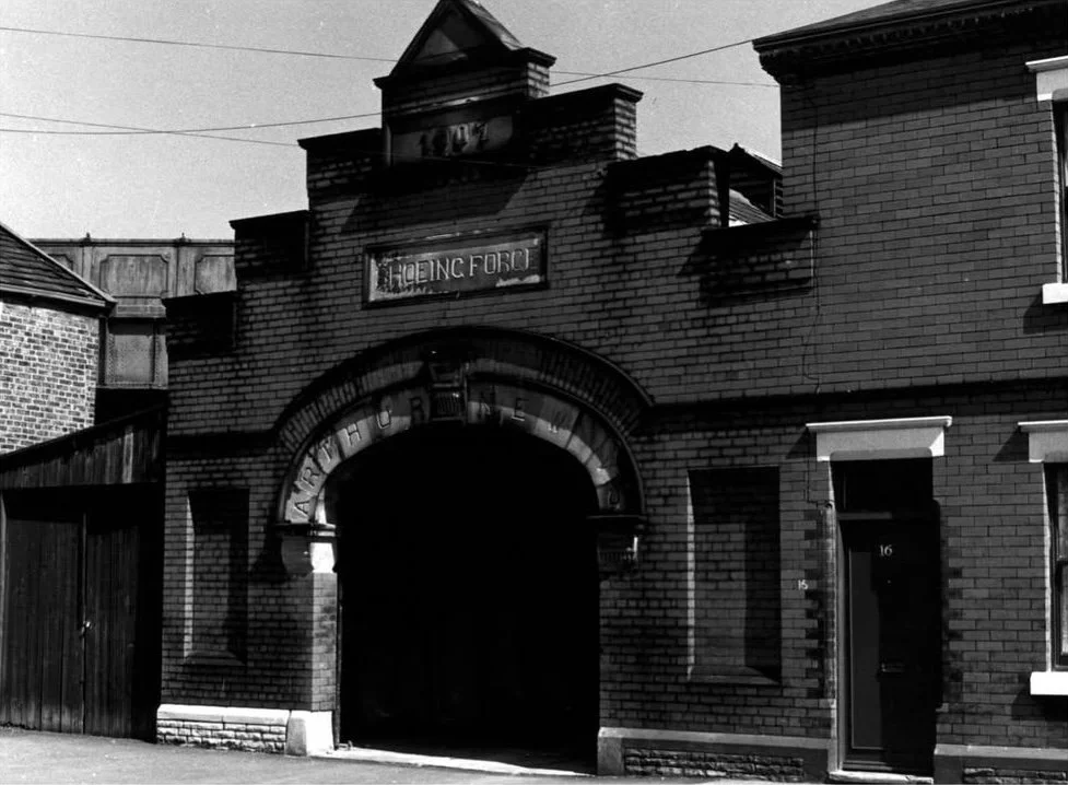 Black and white photo of an old brick archway with a sign above that reads 'Dortachy'. To the right, there is a brick building with a door numbered 16 and a window.
