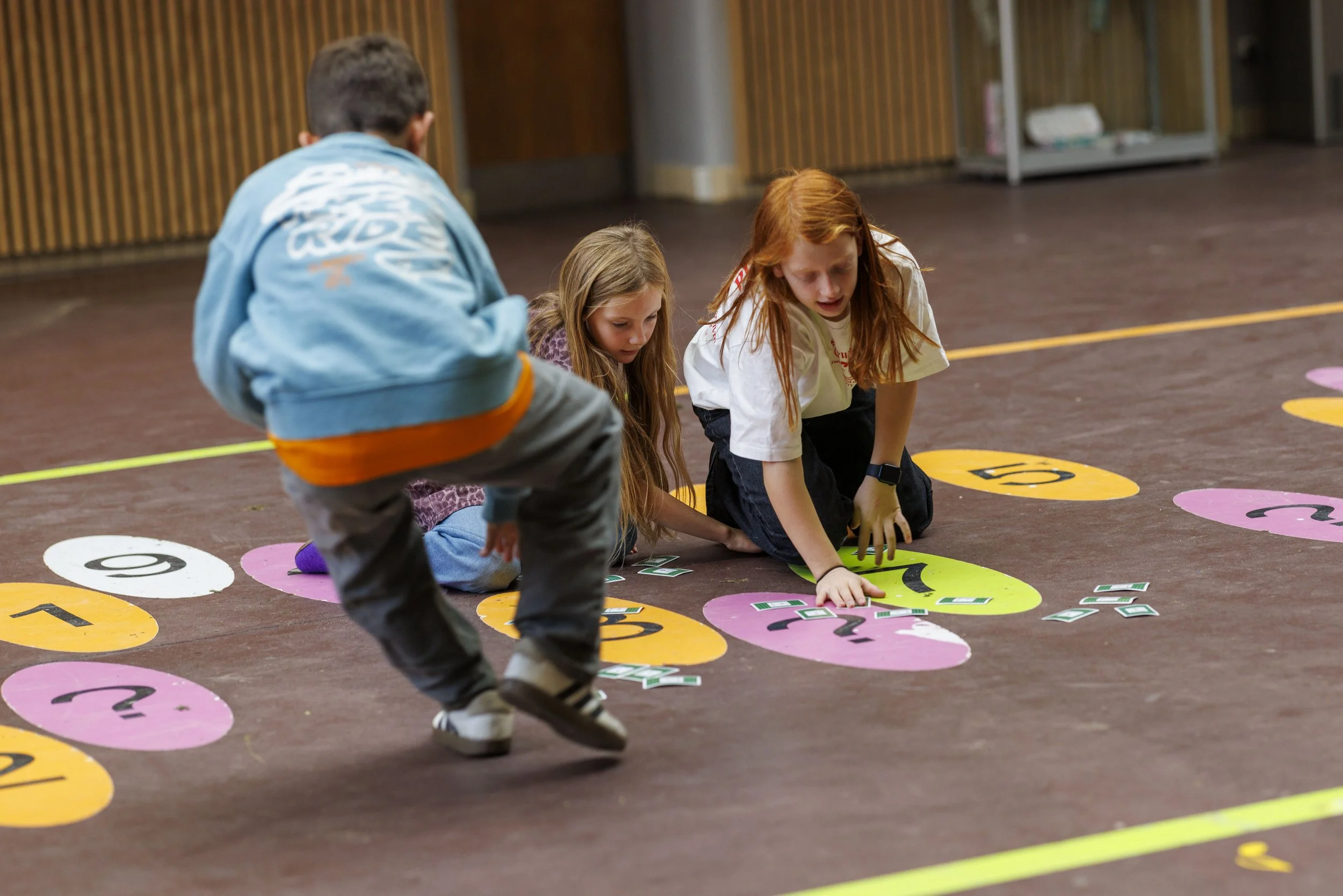 Tre børn leger på gulvet med farvede tal- og skiltepapsirkler med spørgsmålstegn og tal, i et indendørs legemiljø.