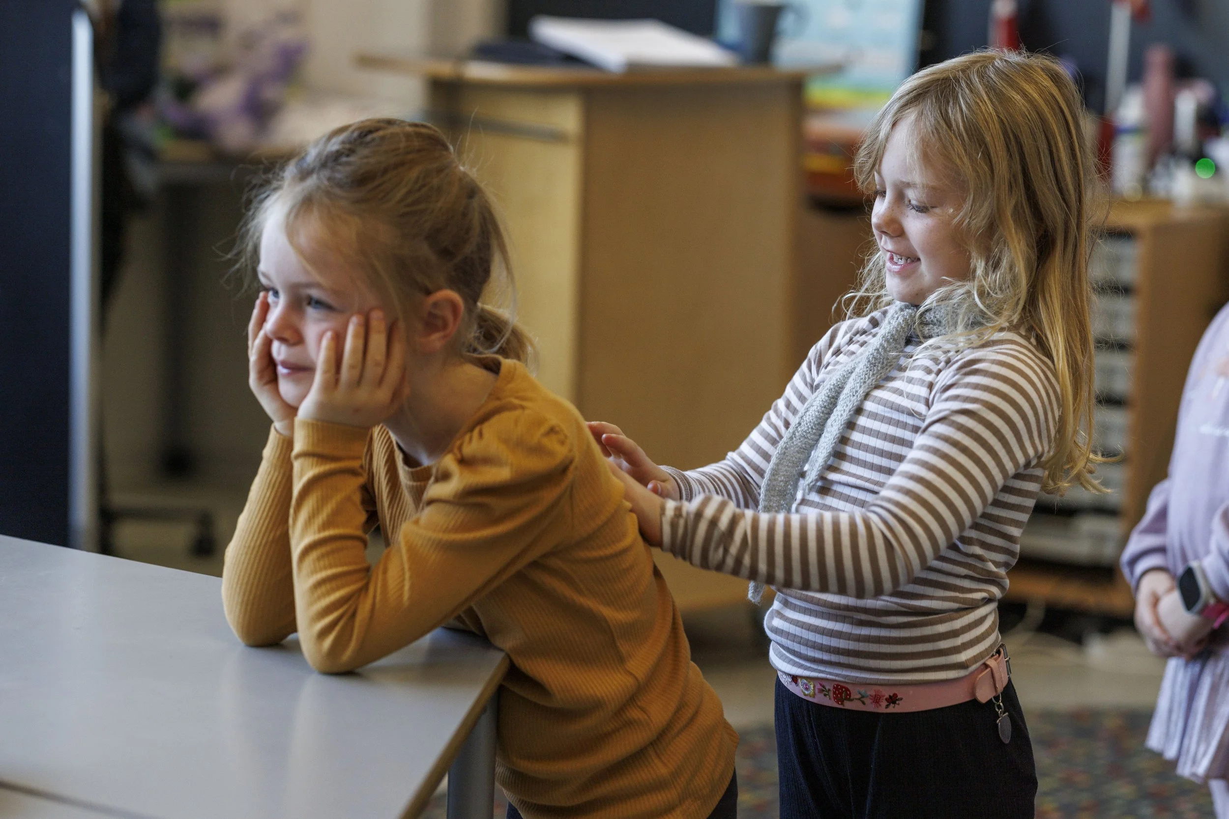 To unge piger der leger i et klasseværelse, hvor den ene sidder med ansigtet i hænderne og den anden leger med hende.