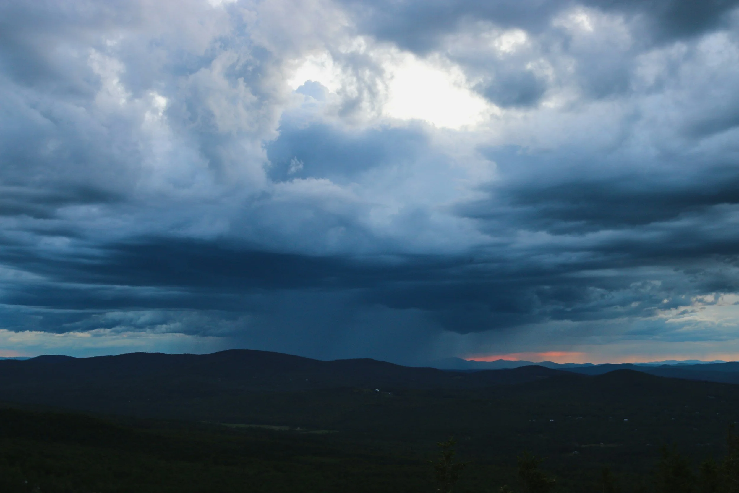 Dark storm clouds over rolling hills at dusk with faint orange horizon.