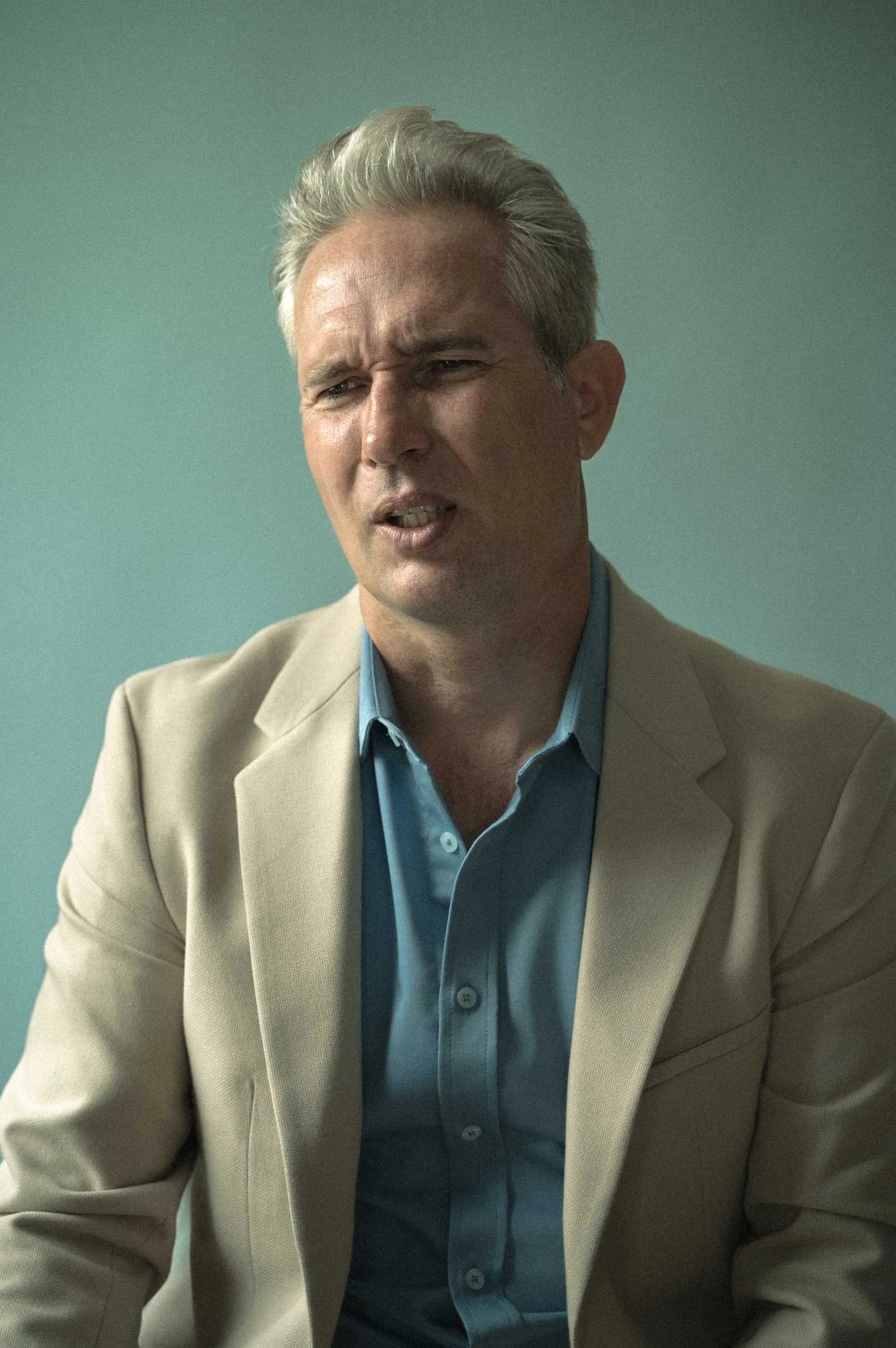Een man met grijs haar in een beige colbert en een blauwe overhemd, zittend en duidelijk in een gesprek.