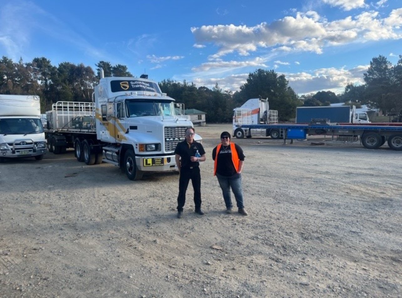 Two people standing in front of large trucks in a gravel lot, with trees and a blue sky with clouds in the background.