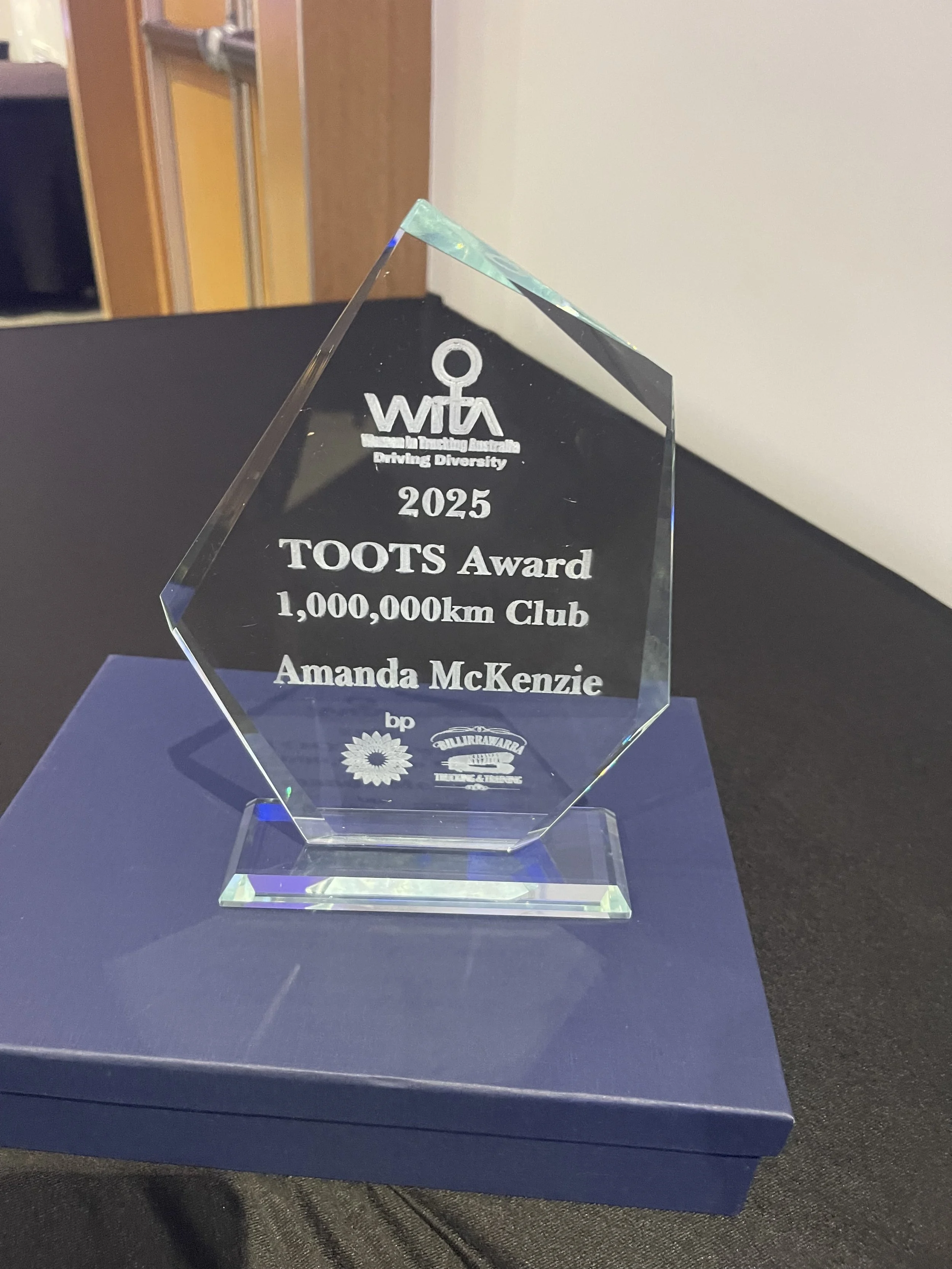 A clear, hexagonal glass award with engraved text. The award is from WIT Australia for driving diversity, dated 2025, for the TOOTS Award for 1,000,000 km Club, awarded to Amanda McKenzie. It features logos of BP and other organizations.