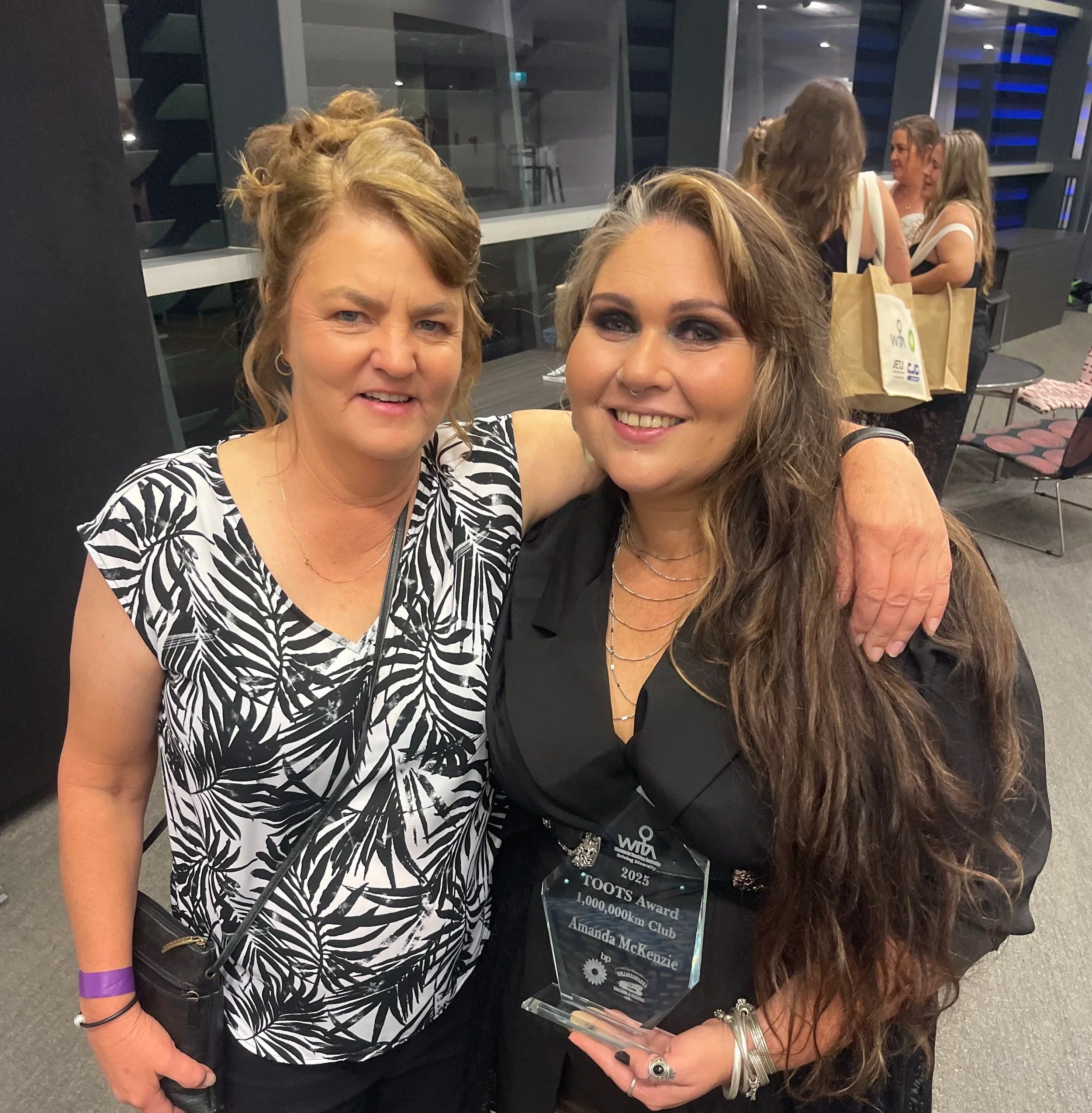 Two women smiling and posing together at an indoor event, one of whom is holding a glass award plaque. In the background, several other women are engaged in conversation and carrying event bags.
