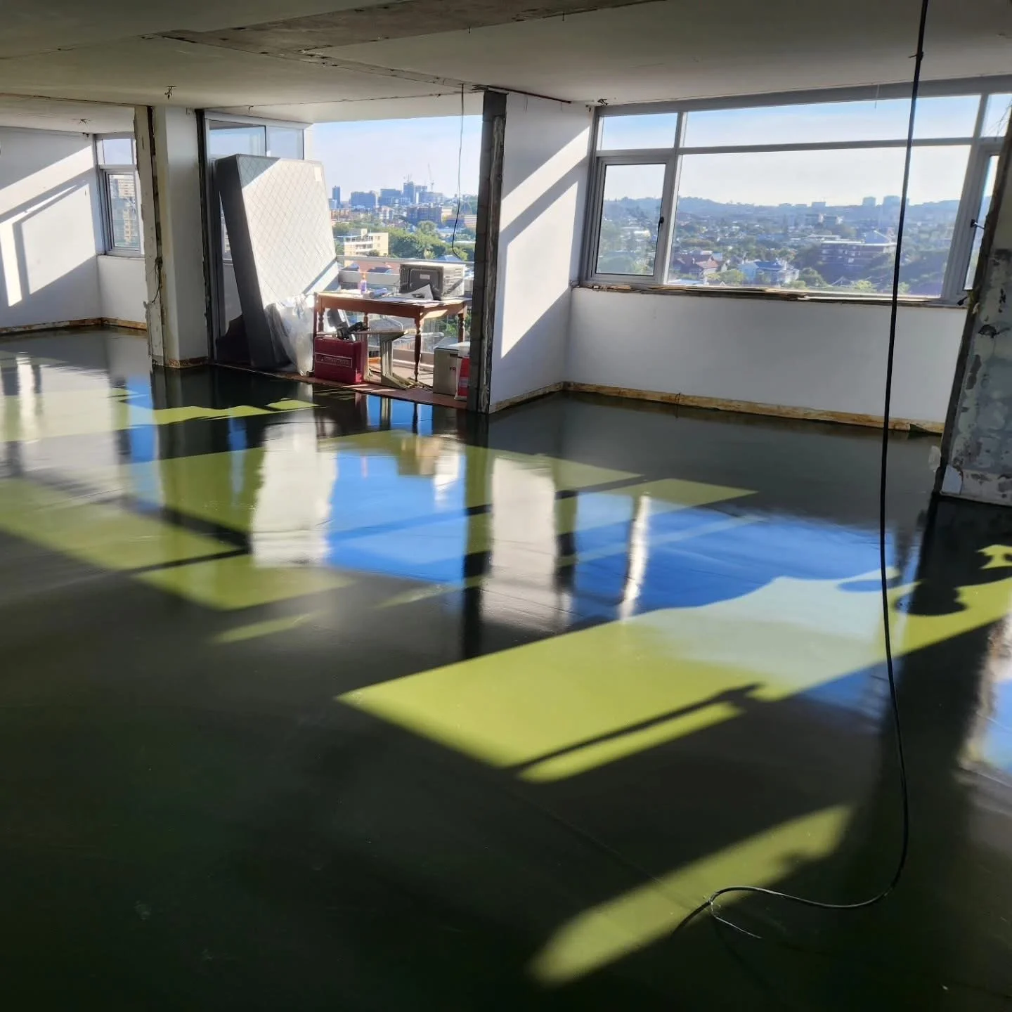Unfinished interior of an apartment with large windows showing a cityscape view, construction materials stored near the window, and a highly polished black floor reflecting sunlight and shadows.