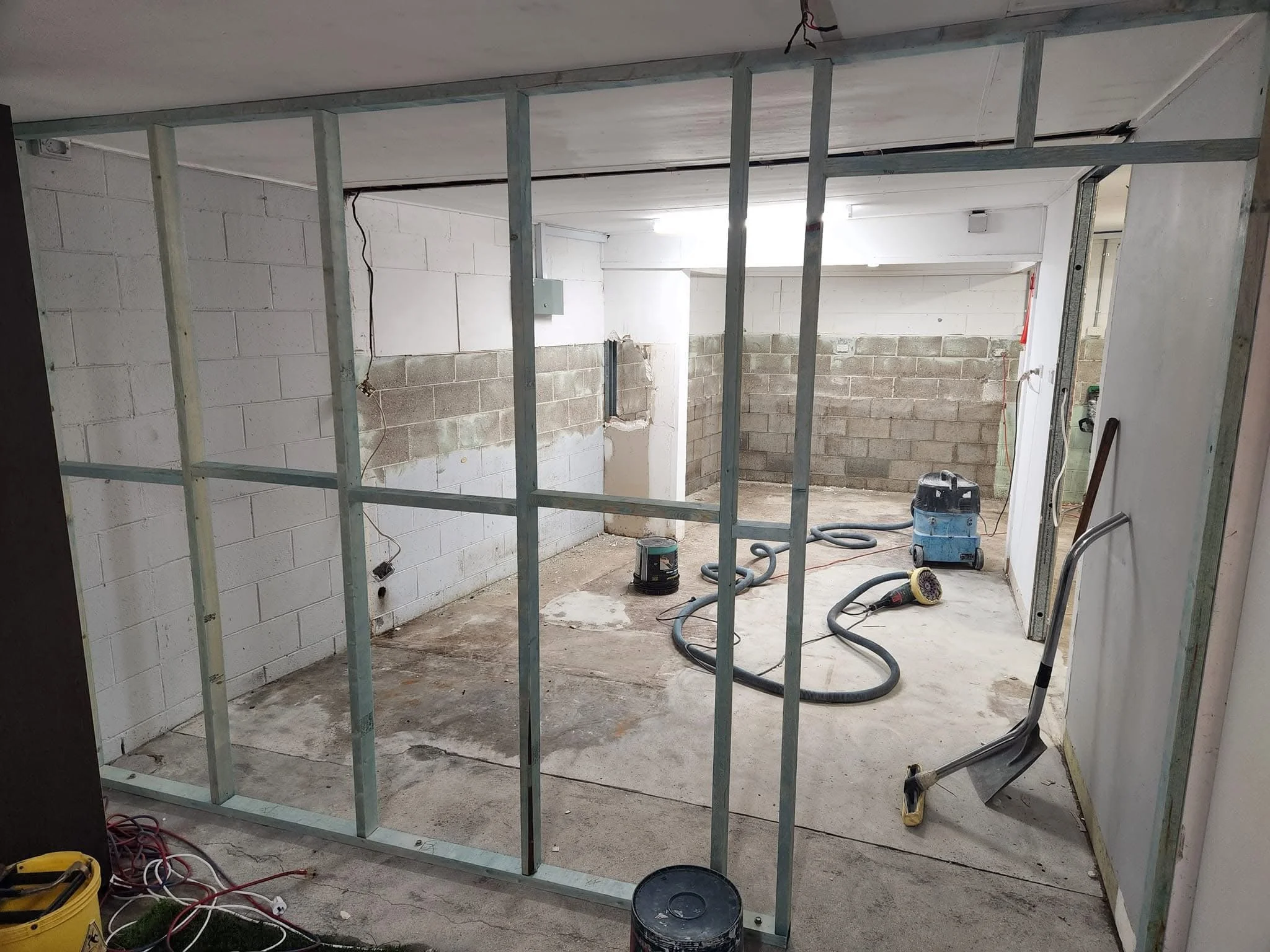 Construction site with metal framing for a new wall in an unfinished room. Tools and hoses are on the floor, cinder block walls, and exposed wiring.