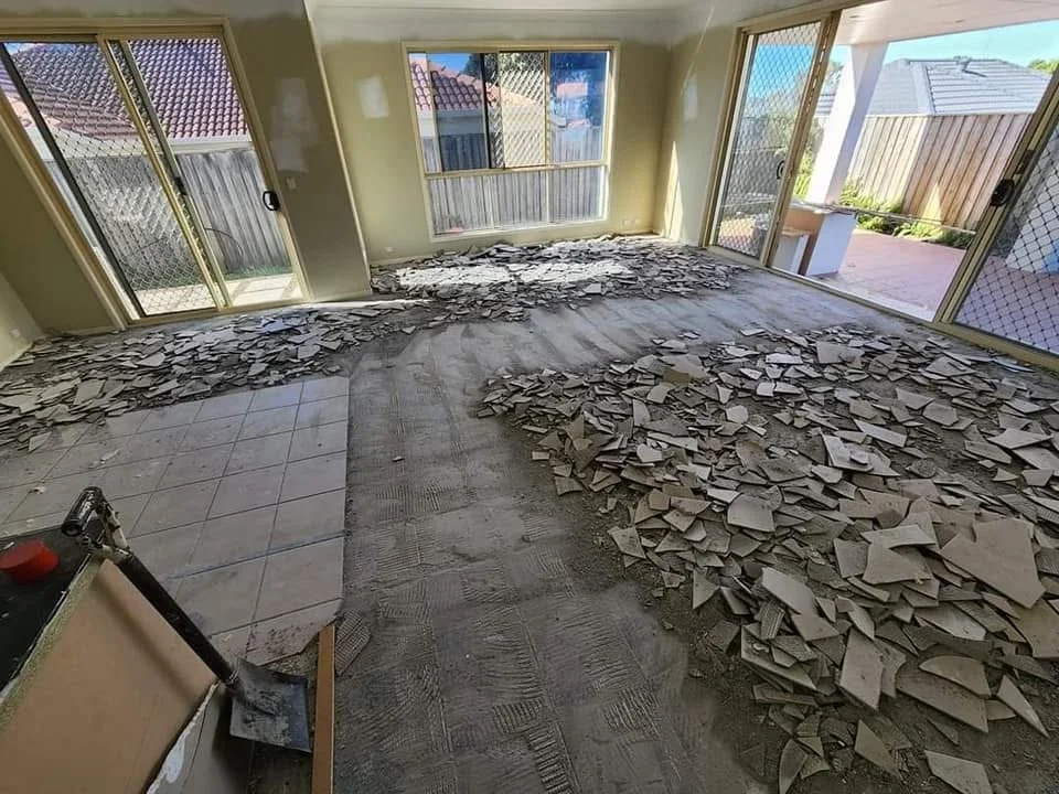 A room under renovation with old tile flooring removed, exposing the subfloor. Piles of broken tile pieces and debris cover the floor. Large windows and sliding glass doors lead to an outdoor patio area.