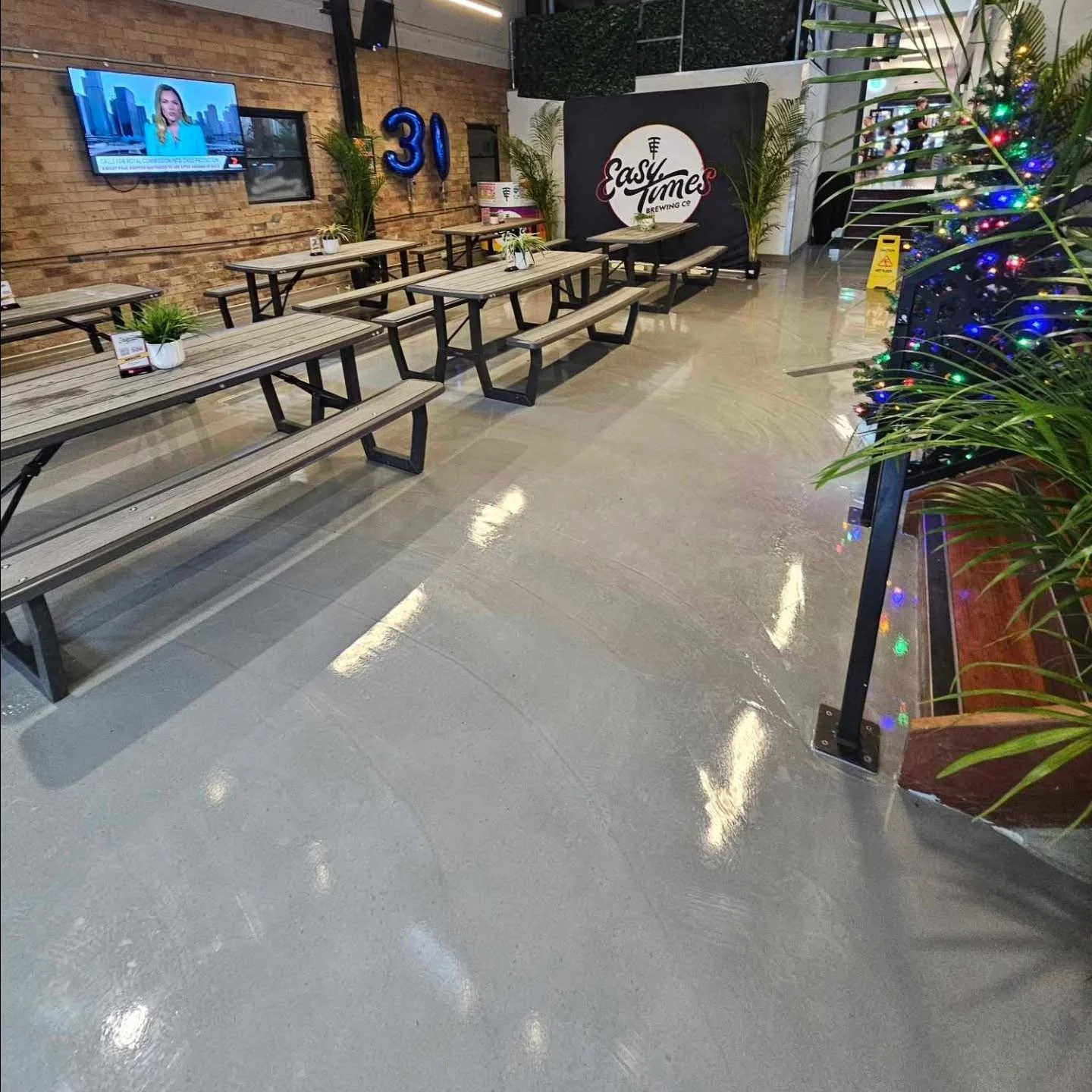 Empty dining area with wooden picnic tables, small potted plants, a television on a brick wall, and a sign for Easy Times Brewing Co. in the background. Holiday decorations include a Christmas tree with colorful lights.