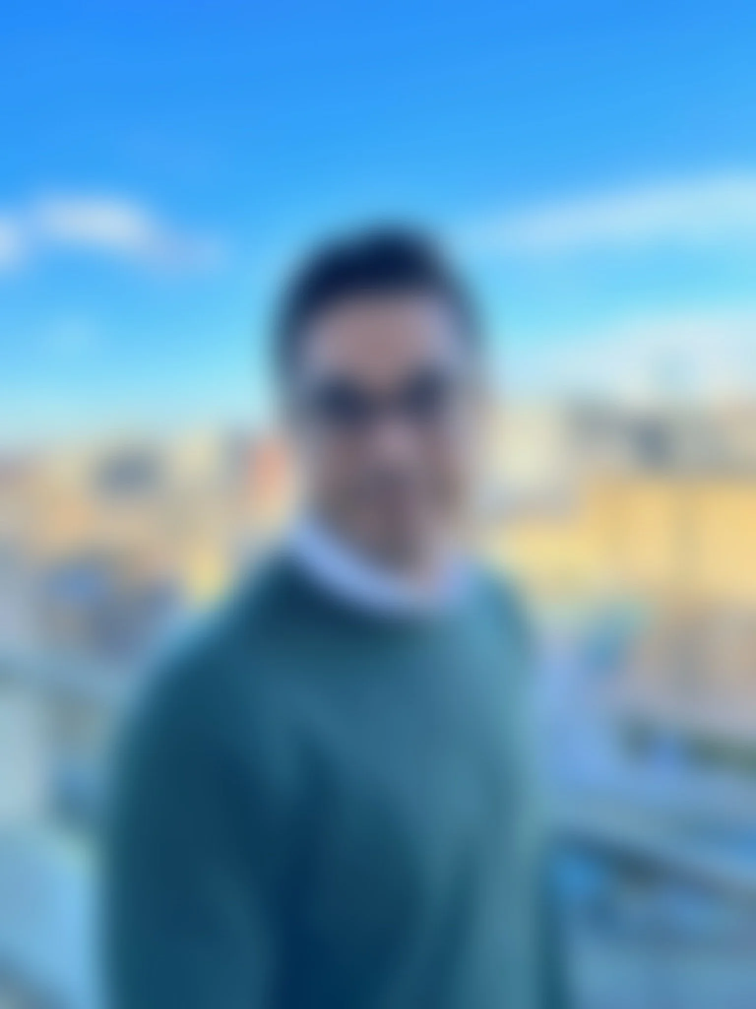 A young man with dark hair, glasses, and a light blue shirt under a dark green sweater standing on a city rooftop with a blue sky and city buildings in the background.