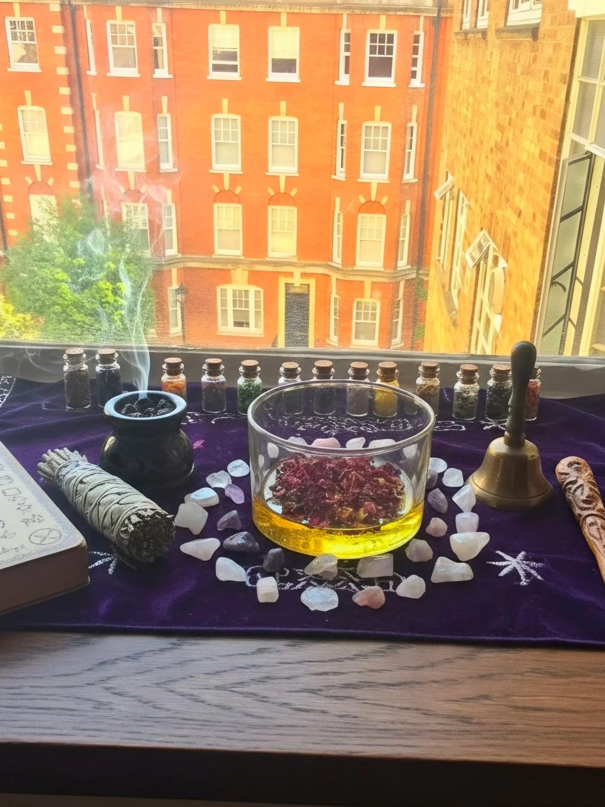A window view with a table set up for spiritual or mystical practices, displaying vials of herbs, a bowl with stones, a candle, a bundle of sage, crystals, and a tarot card, with buildings visible outside the window.