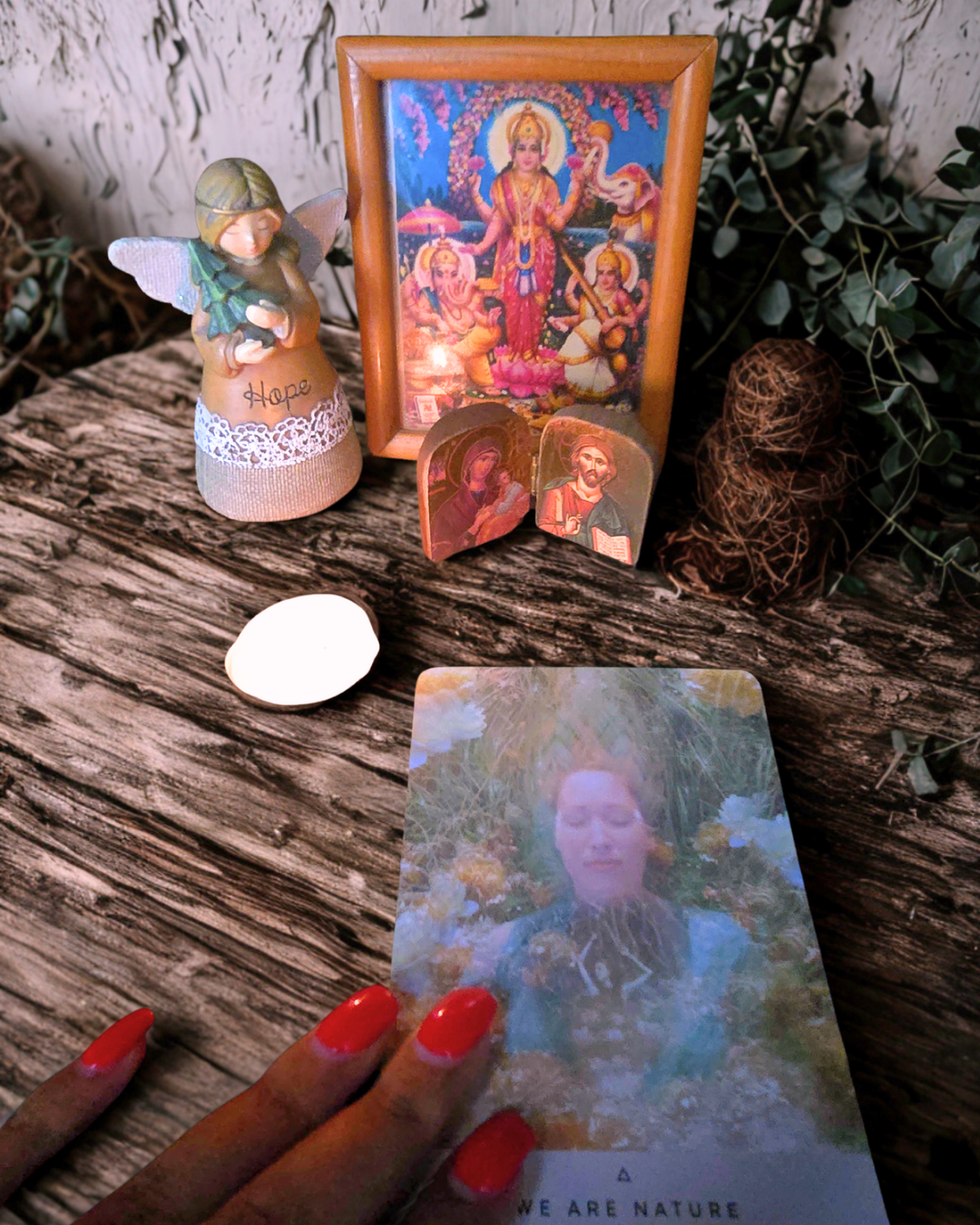A small altar with a framed colorful Hindu goddess picture, a fairy figurine with the word people Hope, two small religious icons, a white stone, a hand with red nails holding a card titled "We Are Nature," some brown string-wrapped objects, and green leaves in the background on a wooden surface.
