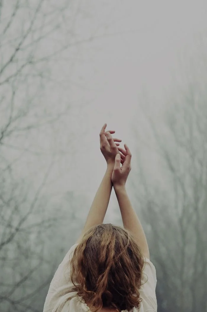 A person with brown curly hair raising their hands against a foggy, leafless tree background.