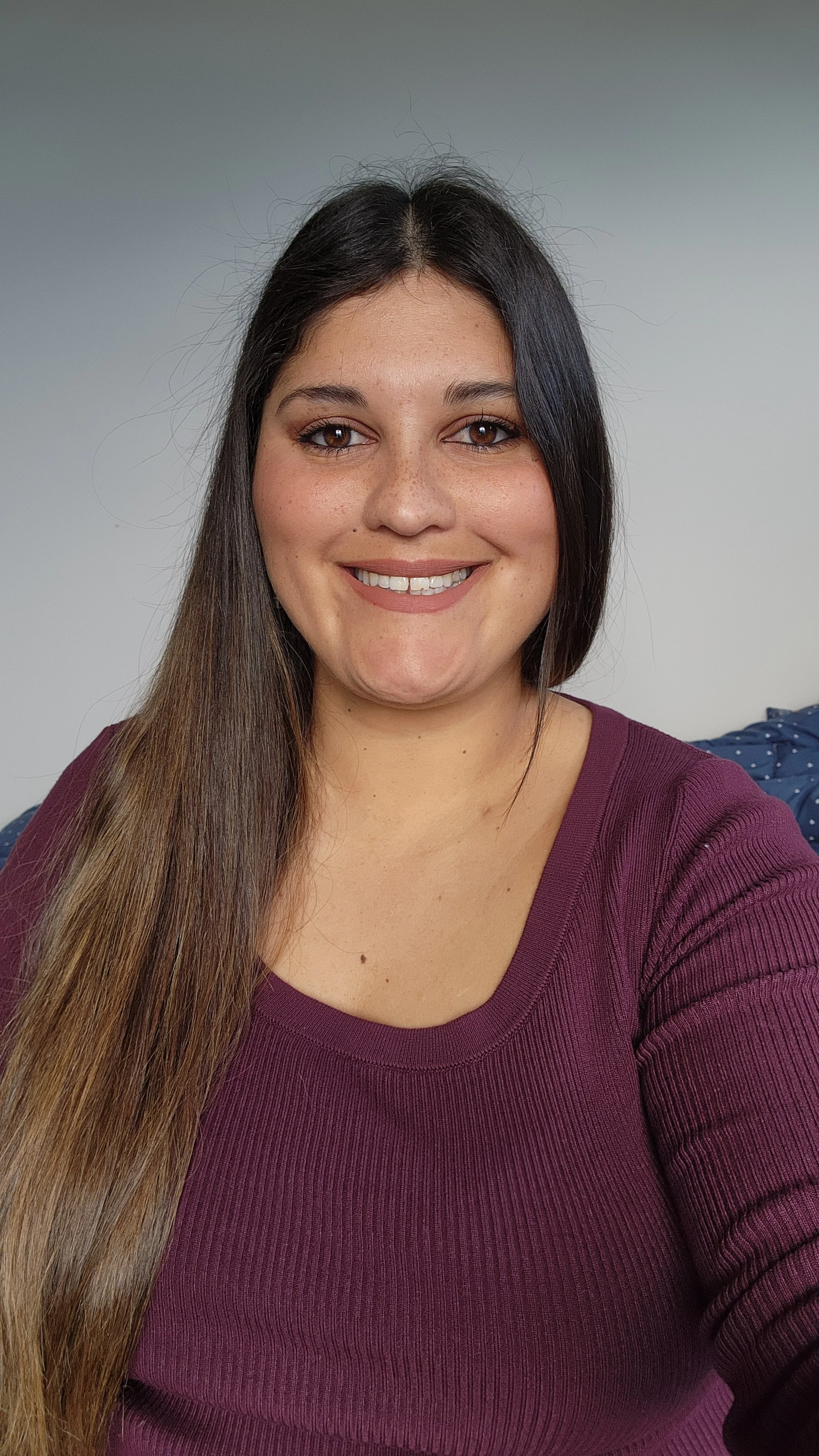 A young woman with long brown hair, smiling and wearing a maroon sweater, sitting on a bed with blue patterned sheets in a room with plain white walls.