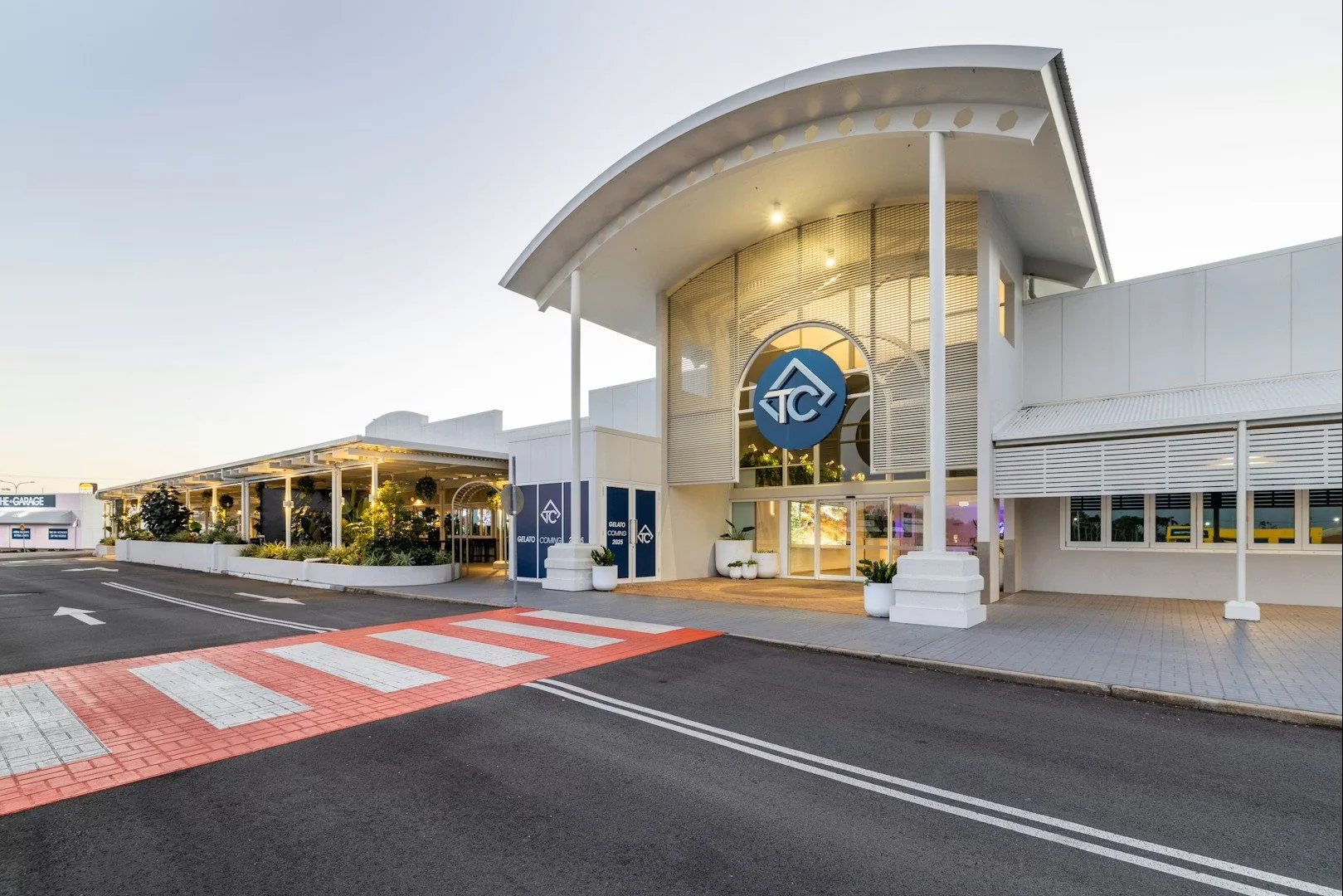 Exterior view of a modern white building with a large circular logo at the entrance, surrounded by potted plants and trees, with a pedestrian crosswalk and road in the foreground.