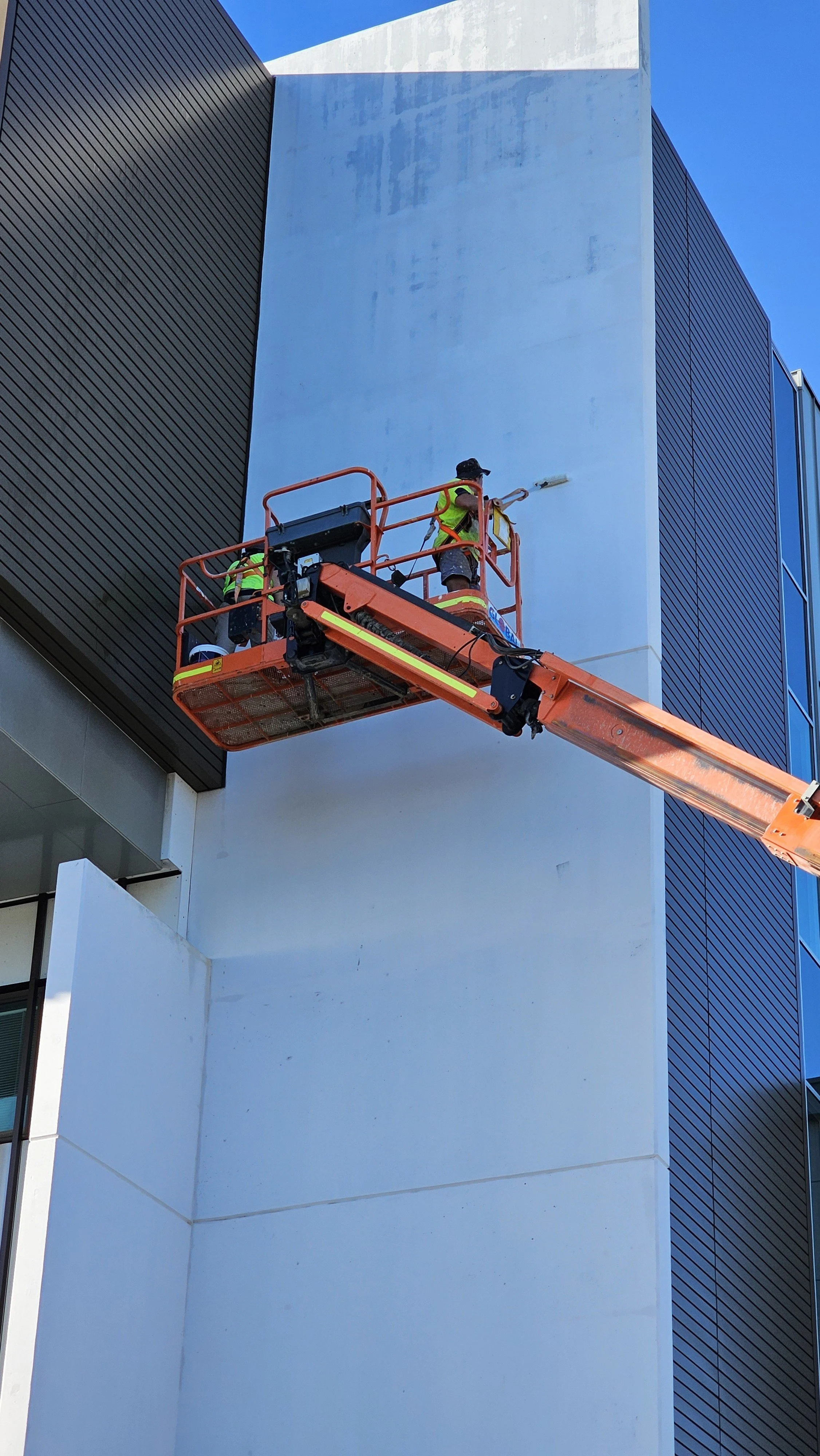 Two workers on an orange lift working on the exterior of a building, painting or cleaning the light gray wall.