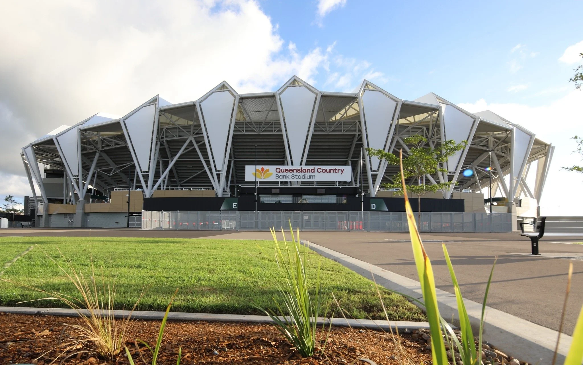 Exterior of Queensland Country Bank Stadium with a sign, a green lawn, and a blue sky with some clouds.