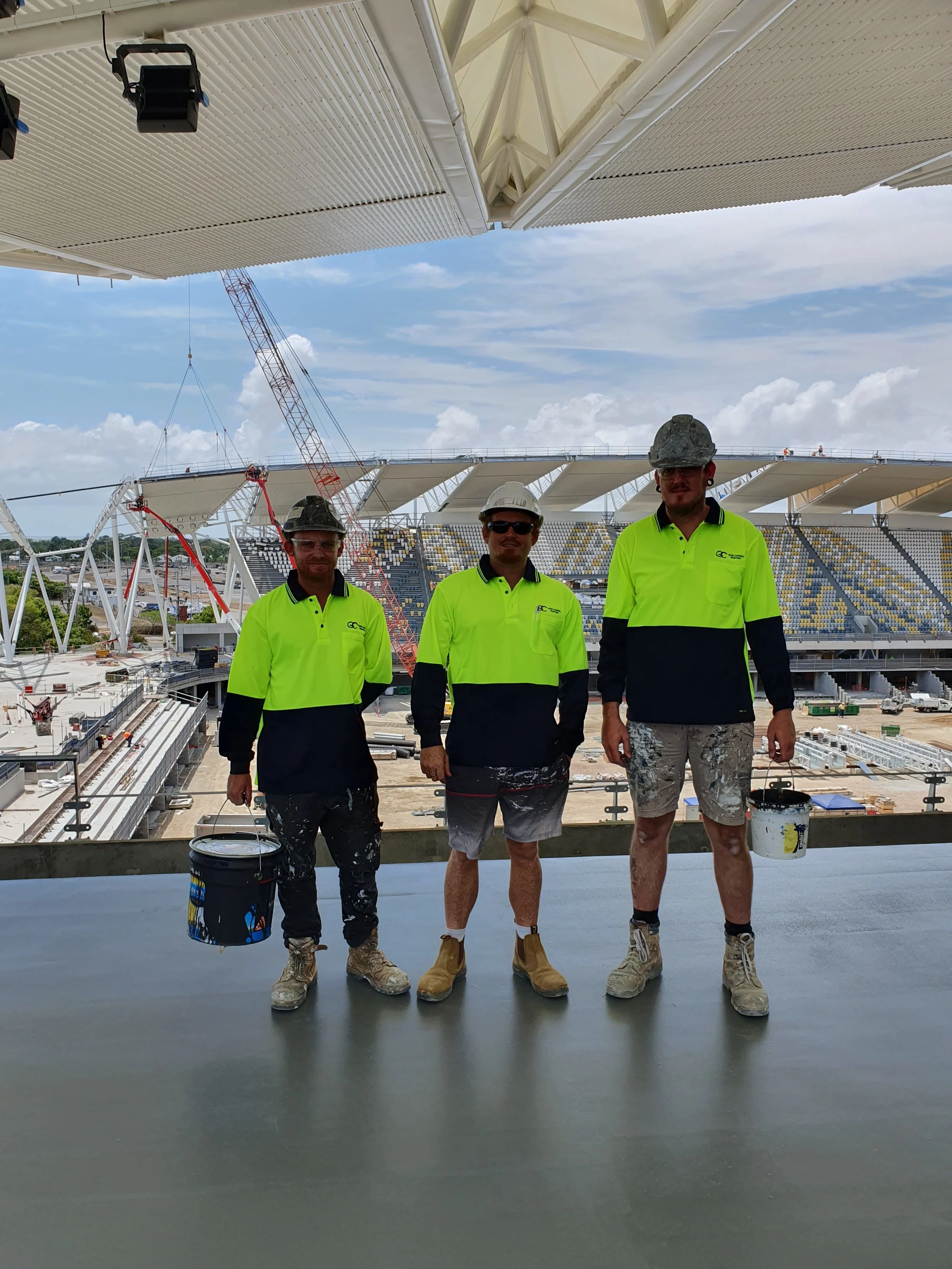 Three construction workers in high-visibility yellow and black shirts, wearing helmets and boots, standing on a construction site with a stadium and cranes in the background.