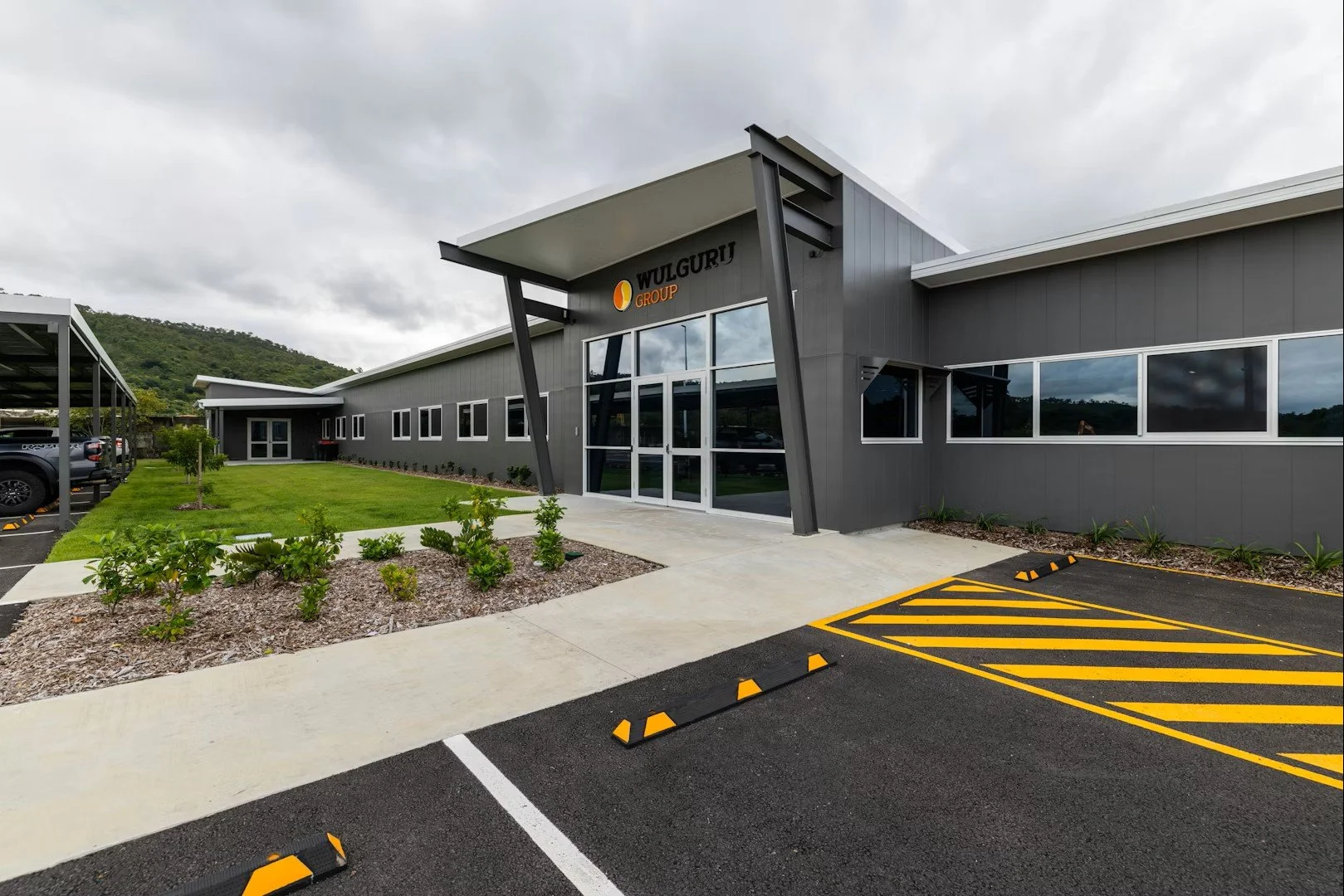 Modern commercial building with the sign 'WULGURI GROUP' above the entrance, surrounded by parking spaces and landscaping, with an overcast sky and green hills in the background.