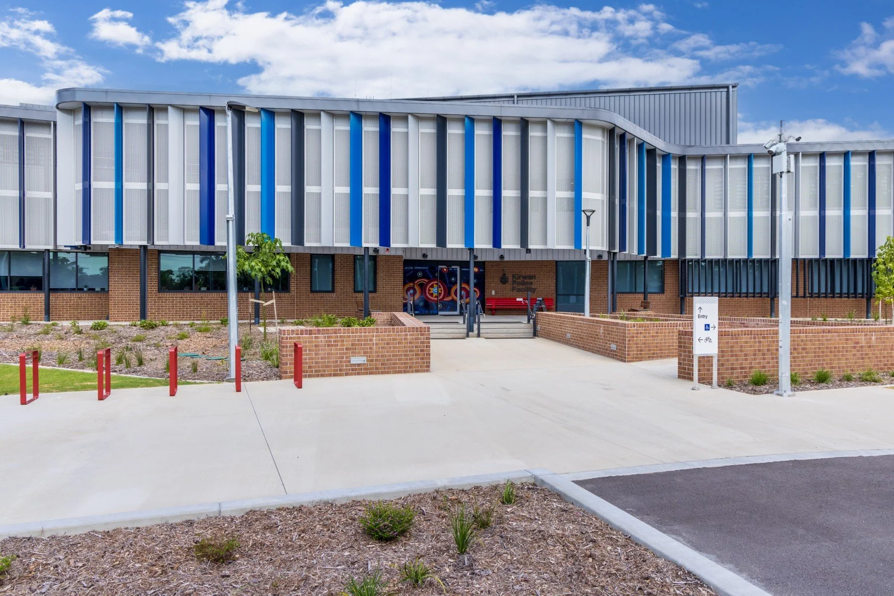 Modern building with brick lower half and metallic upper facade, blue and gray vertical accents, with a wide entrance and ramps, surrounded by landscaped area and parking spaces under a partly cloudy sky.