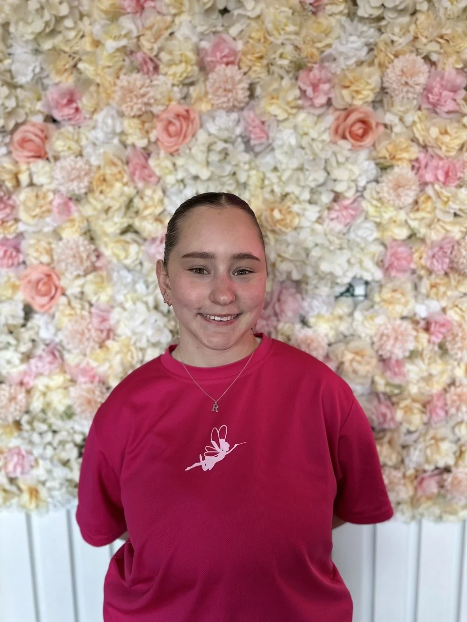 A young girl with light skin and brown hair tied back, smiling and wearing a pink t-shirt with a hummingbird logo, standing in front of a floral backdrop with white, pink, and peach-colored flowers.