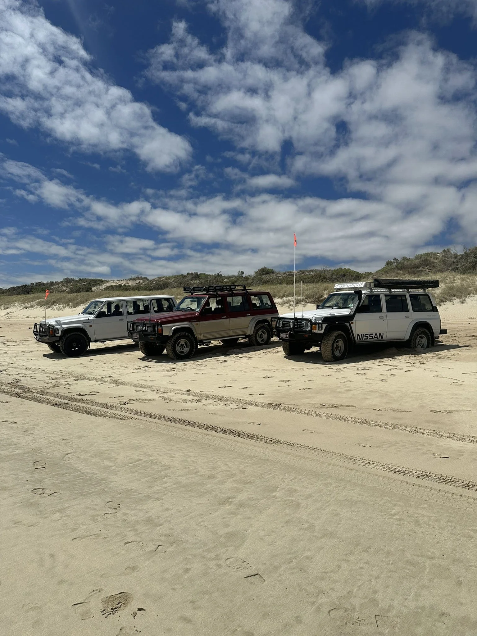 Three off-road vehicles parked on sandy beach with tire tracks, dunes and grassy vegetation in the background, under partly cloudy sky.