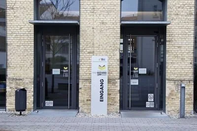 Der Eingang zu einem Gebäude mit einem weißen Steinsäule, die das Wort 'Eingang' auf Deutsch trägt, zwischen zwei Glastüren, umgeben von einer gelben Ziegelwand.