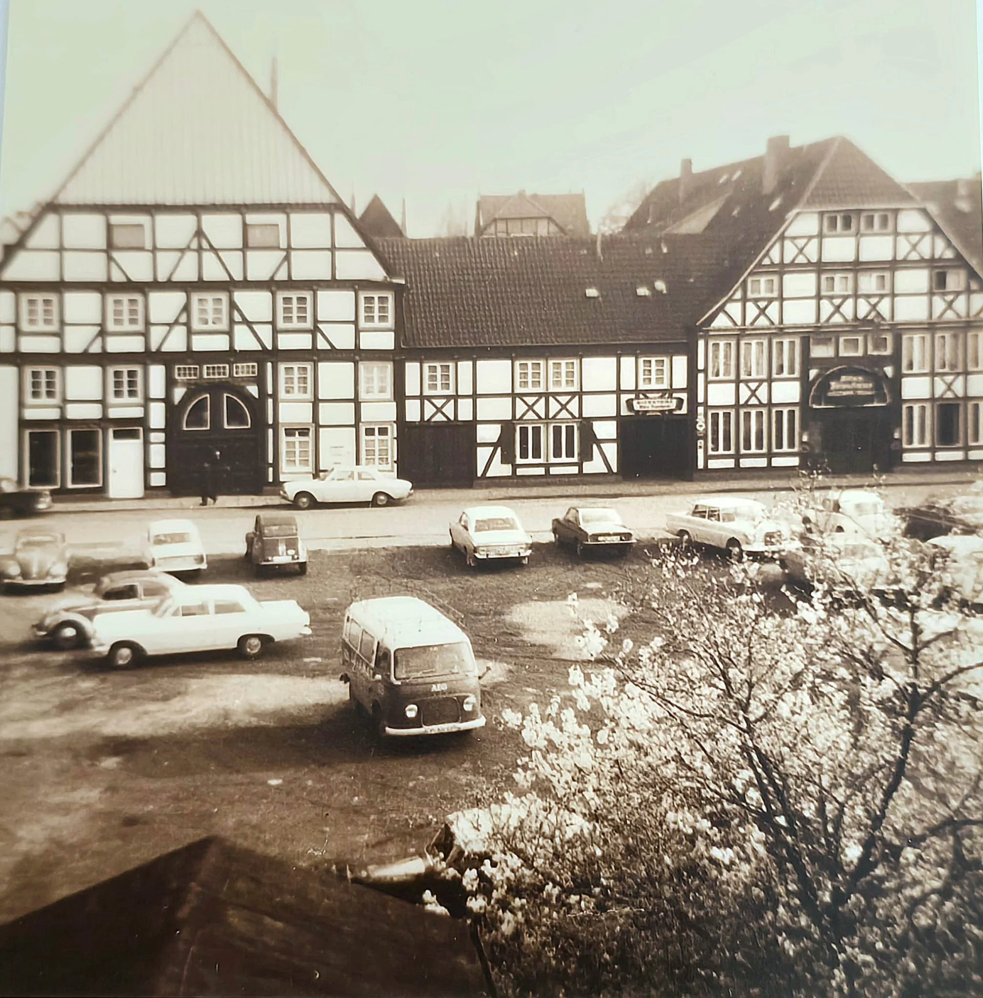 Ein altes Schwarz-Weiß-Foto eines historischen Fachwerkhauses mit mehreren Autos und einem Garten im Vordergrund.