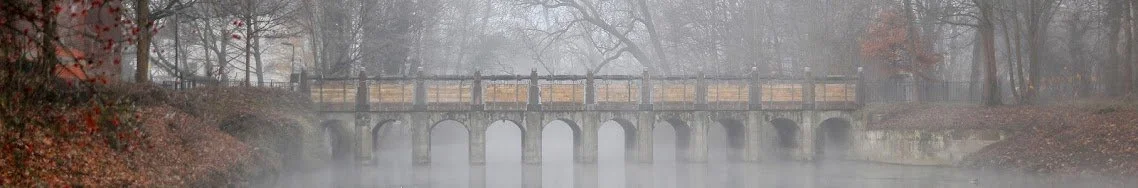 Ein Fluss mit einem kleinen Brücke aus Steinen, umgeben von Bäumen im Nebel.