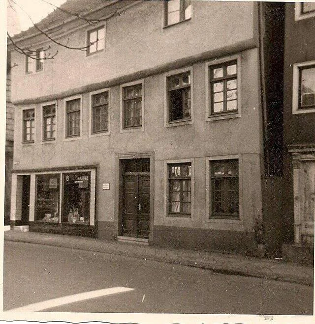 Ein altes, mehrstöckiges Gebäude mit mehreren Fenstern, einige geöffnet, und einer Tür in einer ruhigen Stadtstraße. Es gibt auch Geschäfte im Erdgeschoss.