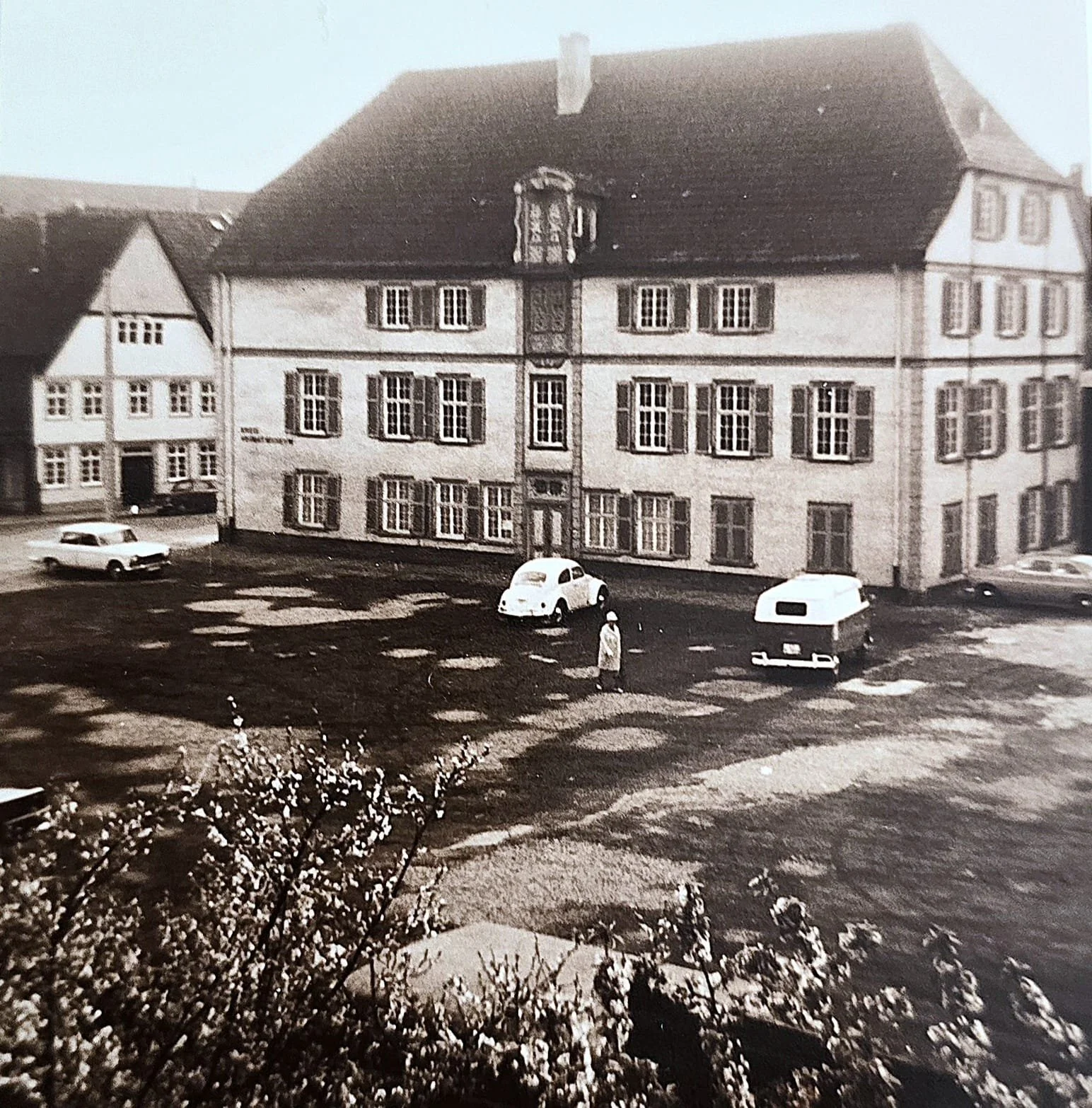 Schwarz-Weiß-Foto eines Gebäudes mit mehreren Fenstern, einem Uhrenturm in der Mitte, einem Auto, einem Wohnwagen und einer Person im Vordergrund, umgeben von Bäumen.