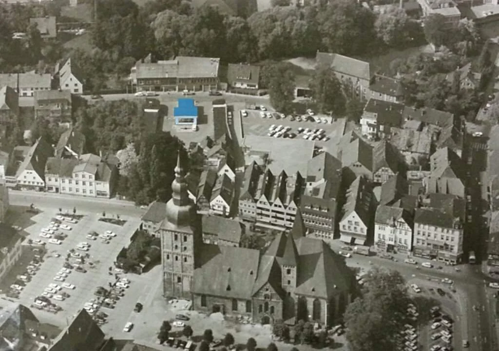 Luftaufnahme einer vielfältigen mittelalterlichen Stadt mit Kirche im Zentrum, umgeben von Wohnhäusern, einem Parkplatz und Grünflächen.