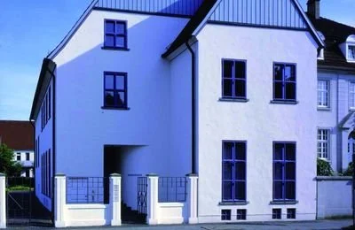 Ein modernes, weißes Mehrfamilienhaus mit blauen Fenstern und Dach, umgeben von einer kleinen weißen Mauer mit Metallzaun.