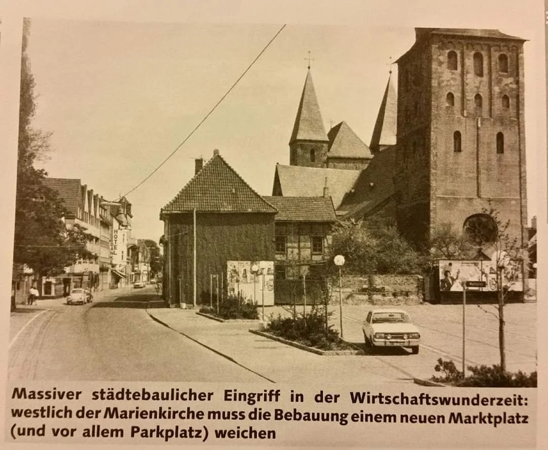 Historisches Foto einer Stadtstraße mit Kirche im Hintergrund, parkenden Autos und mehreren Gebäuden, darunter ein Hotel. Die Straße ist relativ leer, mit wenigen Menschen unterwegs.