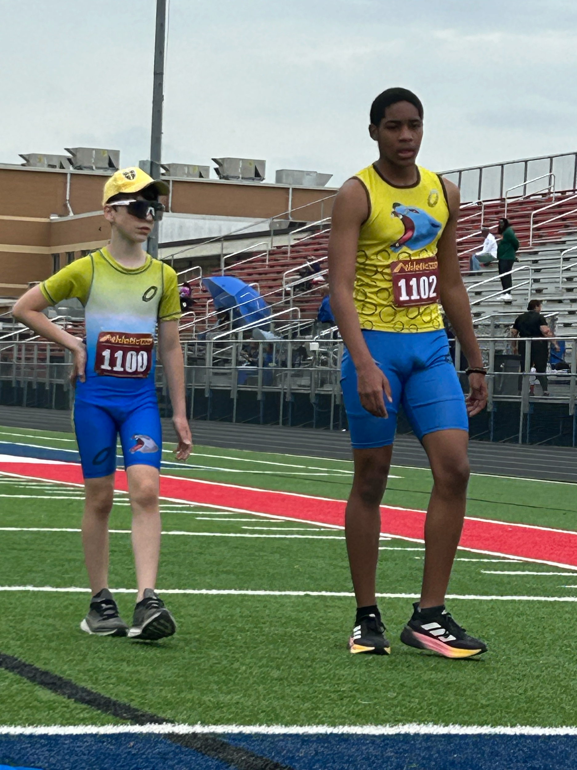 Two young male athletes wearing colorful running gear and bib numbers 1100 and 1102 on a track field, preparing for a race as spectators and officials are present in the background.