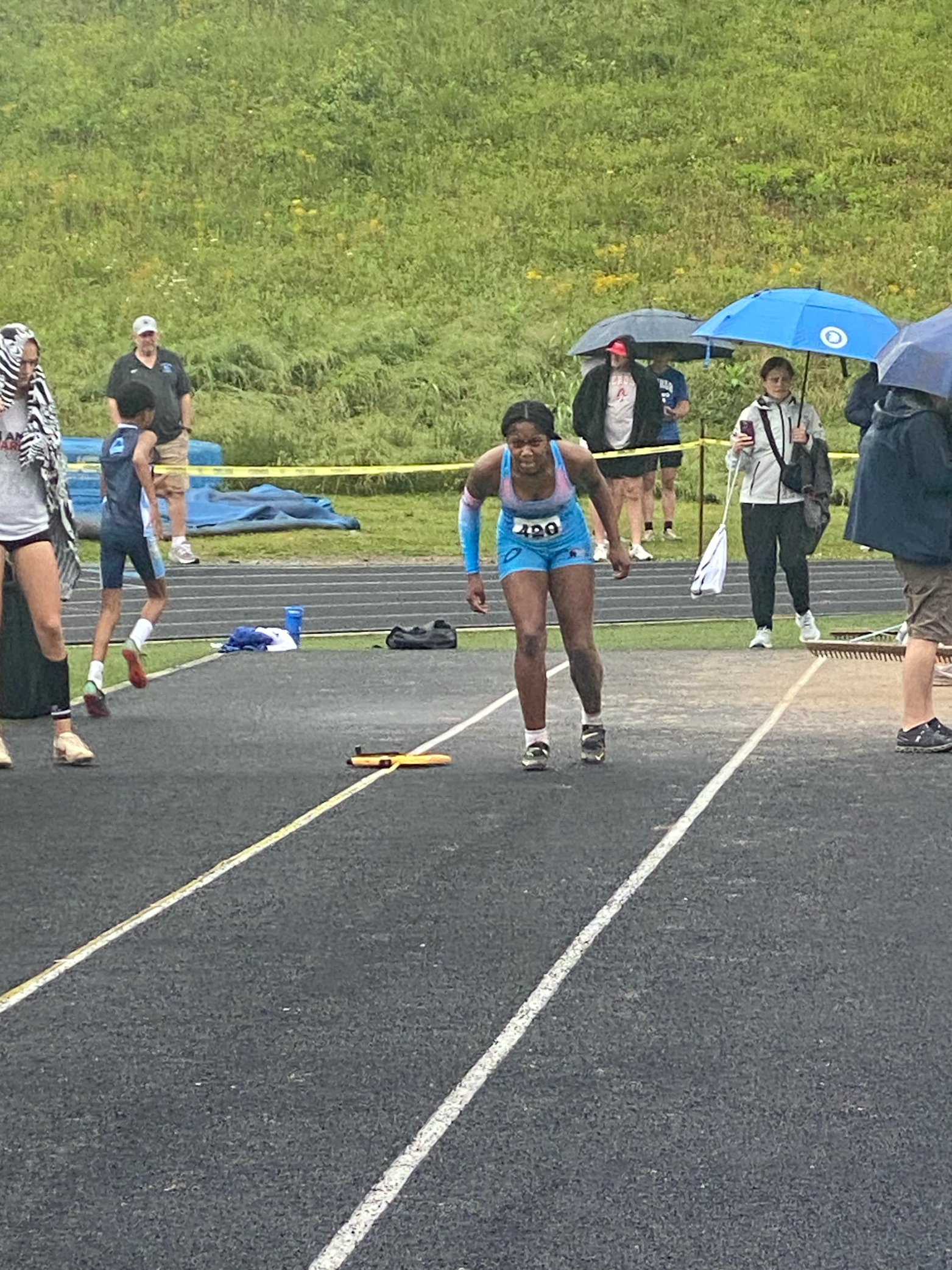 A female athlete is finishing on the triple jump runway, starting her approach track with wet conditions, surrounded by spectators holding umbrellas.