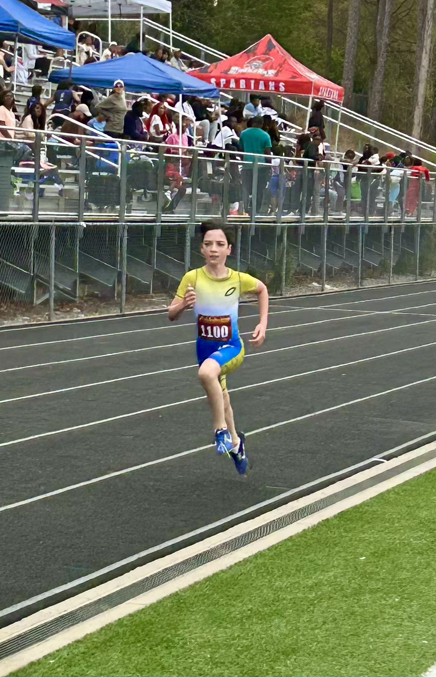 A young boy running on a track during a race, with spectators and tents in the background.