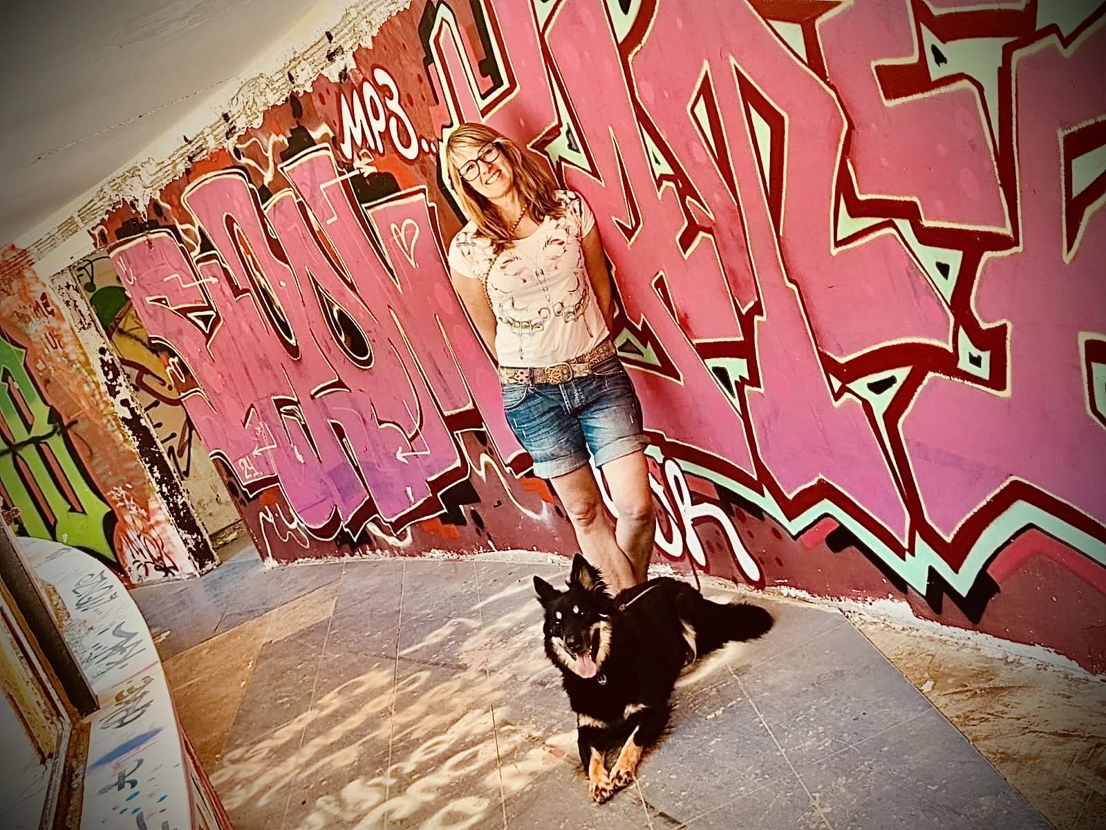 Eine Frau mit Brille und Kurzhaarfrisur steht vor einer bunten Graffiti-Wand, neben ihr liegt ein schwarzer Hund mit braunen Abzeichen, beide schauen zur Kamera, die Hund hat den Mund offen und sieht freundlich aus.
