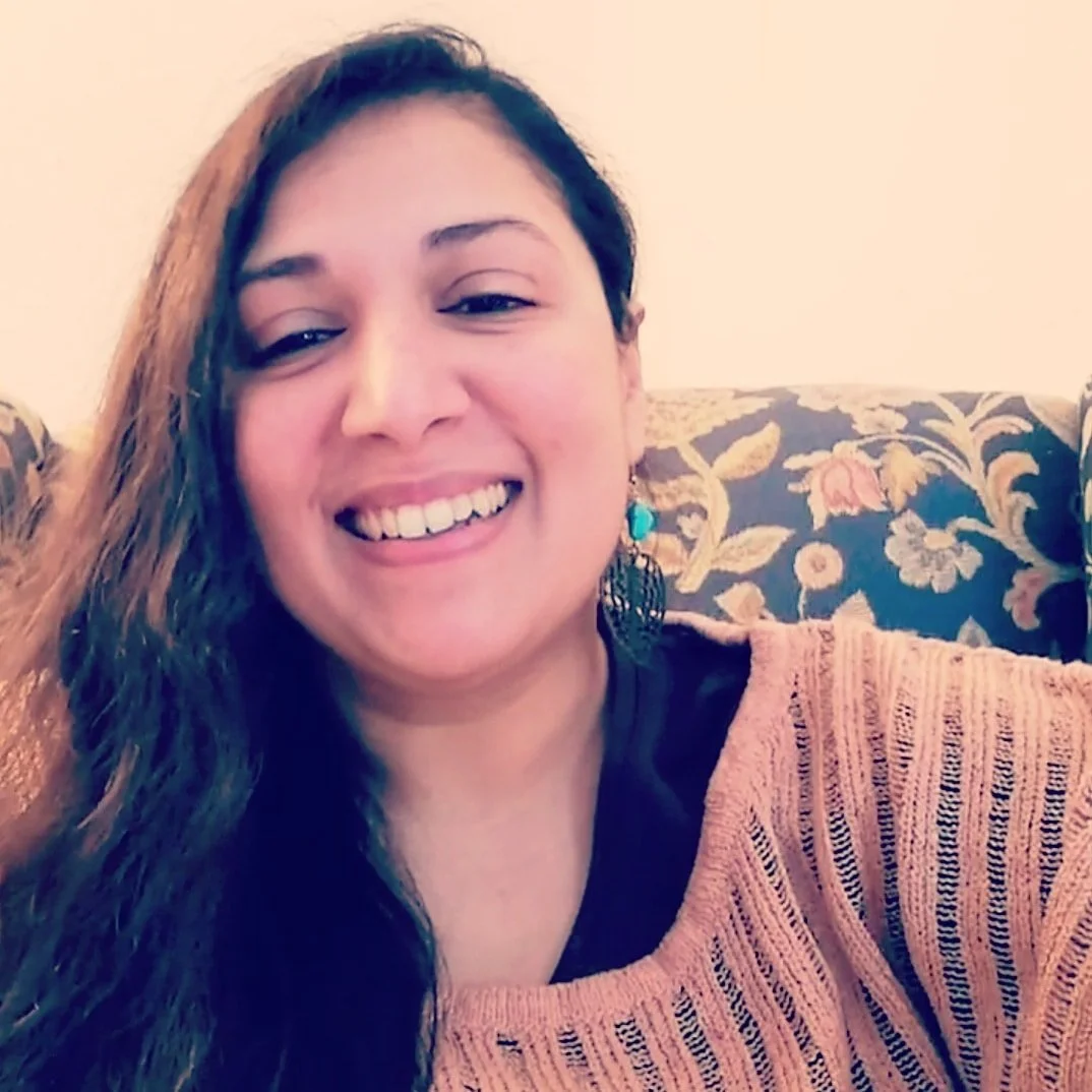 Smiling woman with long brown hair, wearing turquoise earring and a burnt orange sweater, sitting on a floral patterned couch.